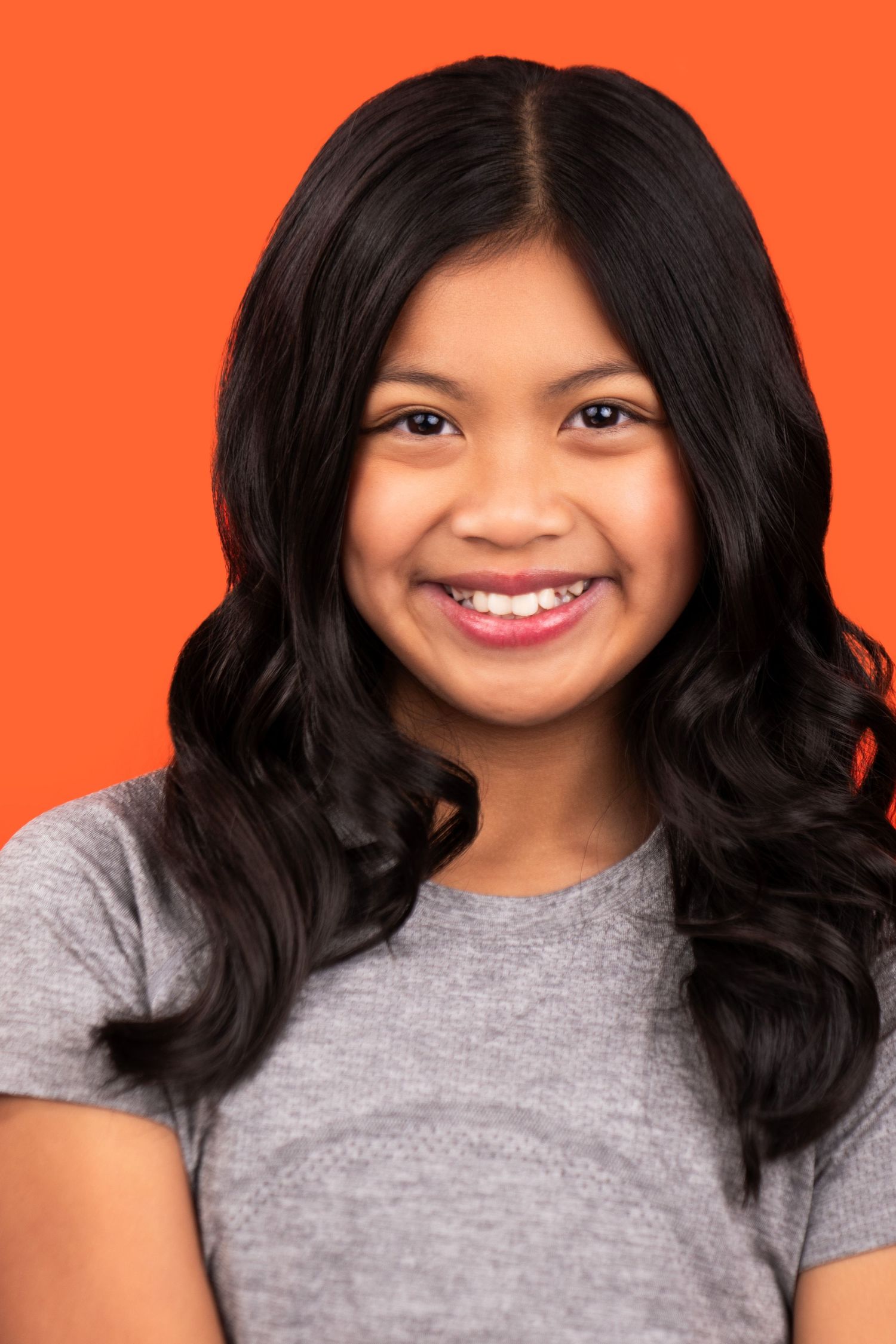 Smiling young woman with long dark wavy hair wearing a grey t-shirt against an orange background.