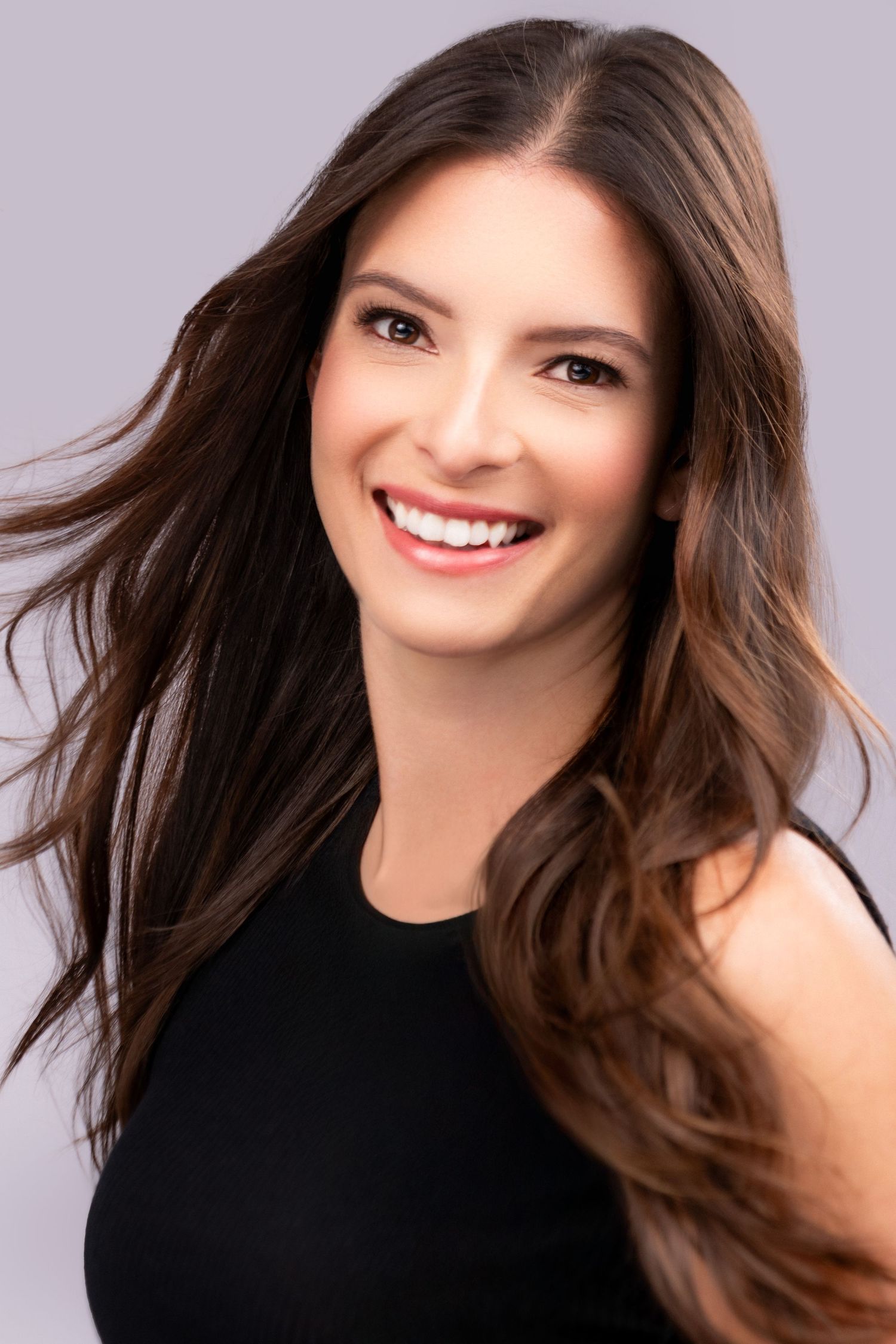 Professional portrait of someone with flowing dark brown hair and bright smile wearing a black sleeveless top against purple background.