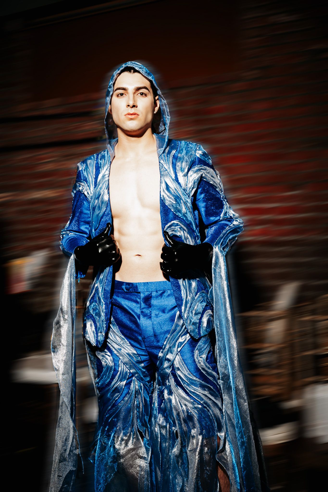 Model wearing shimmering blue artistic fashion ensemble with matching hooded coat on dark runway.