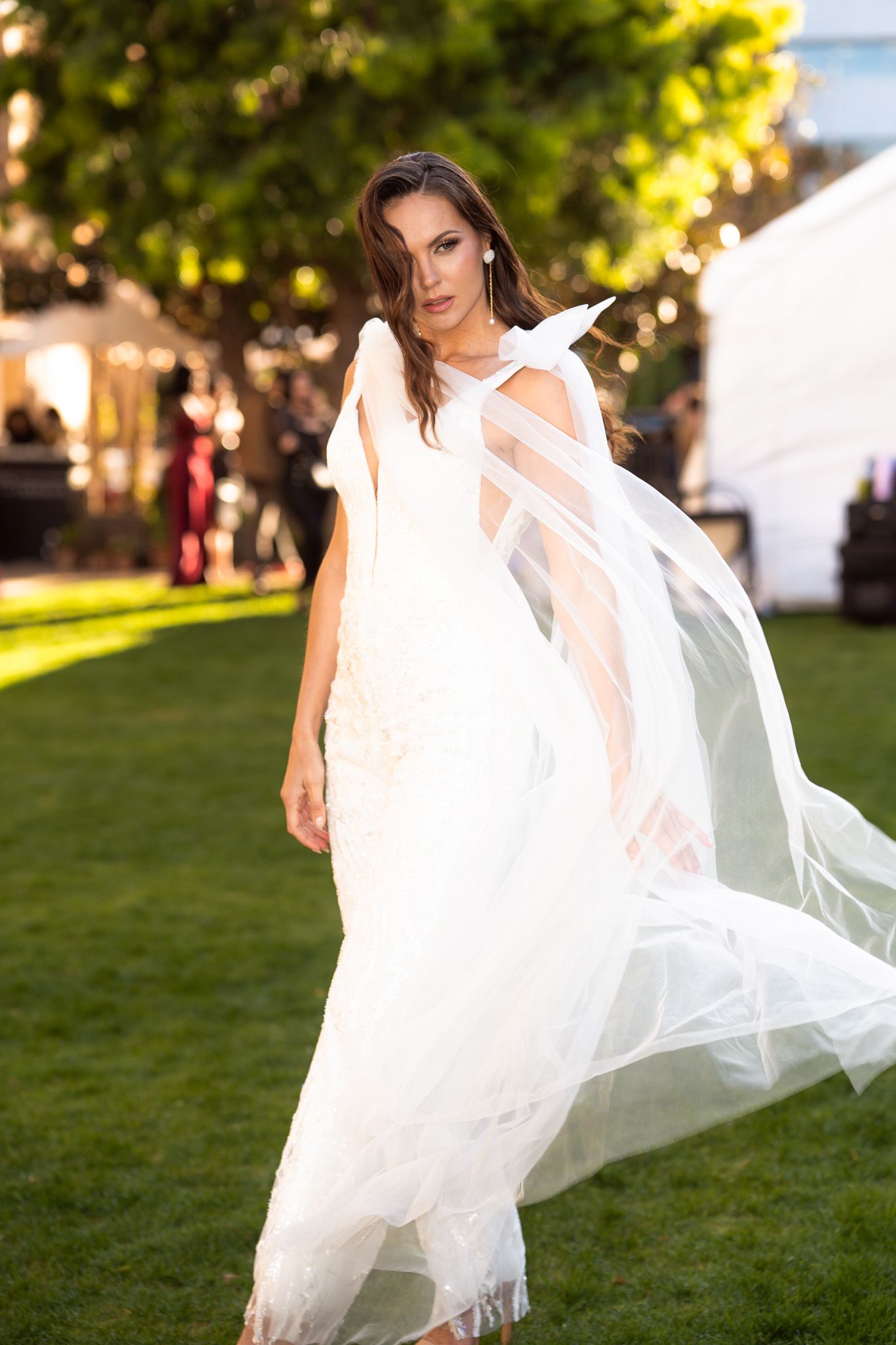 A person in a flowing white wedding dress with sheer fabric details poses on a grassy lawn at an outdoor event.