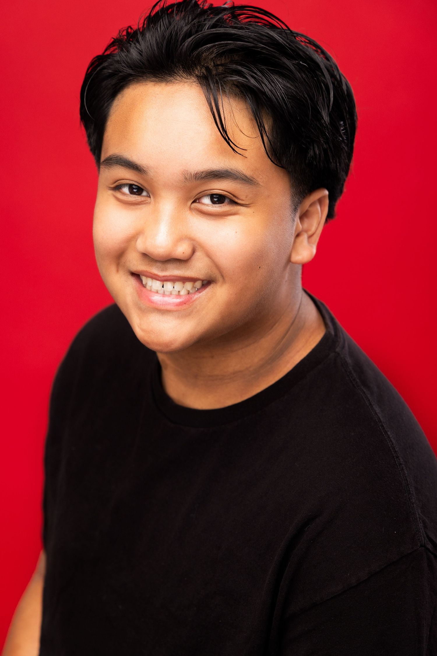 Portrait headshot of someone with black hair wearing a black shirt against a bright red background, smiling warmly at camera.