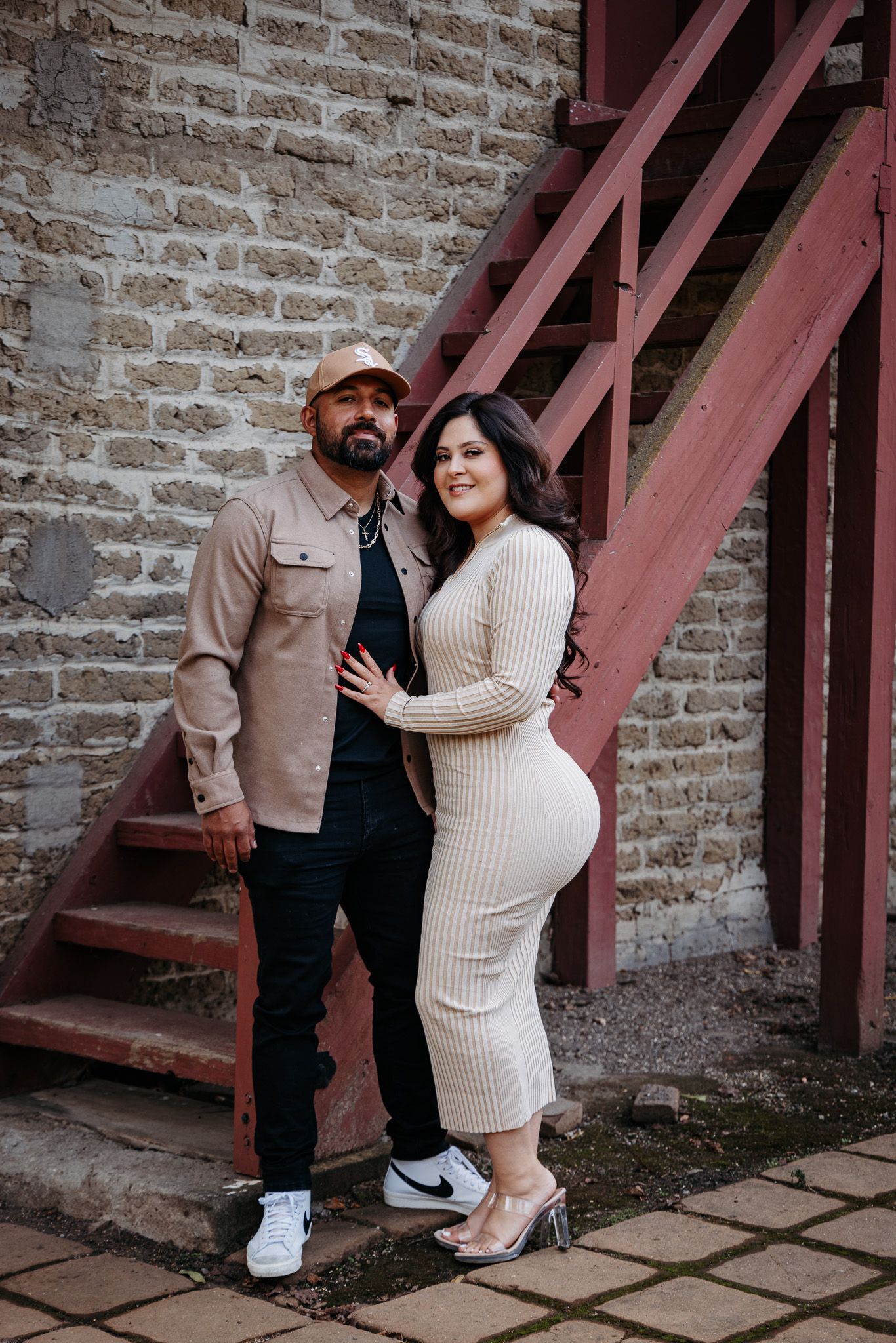 A couple poses together on red metal stairs against a rustic brick wall for a stylish urban portrait session.