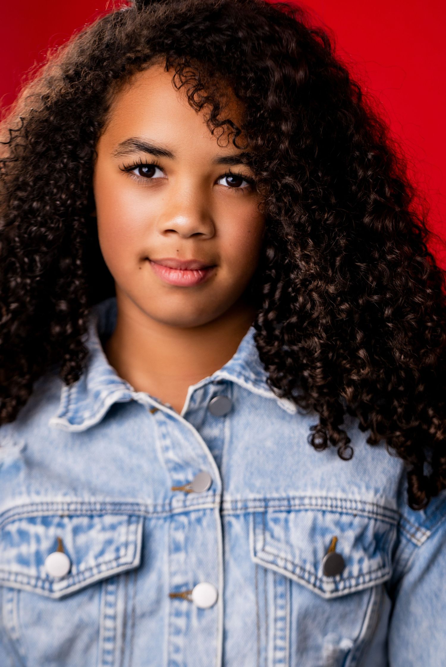 Portrait of someone with long curly dark hair wearing a denim jacket against a red background.