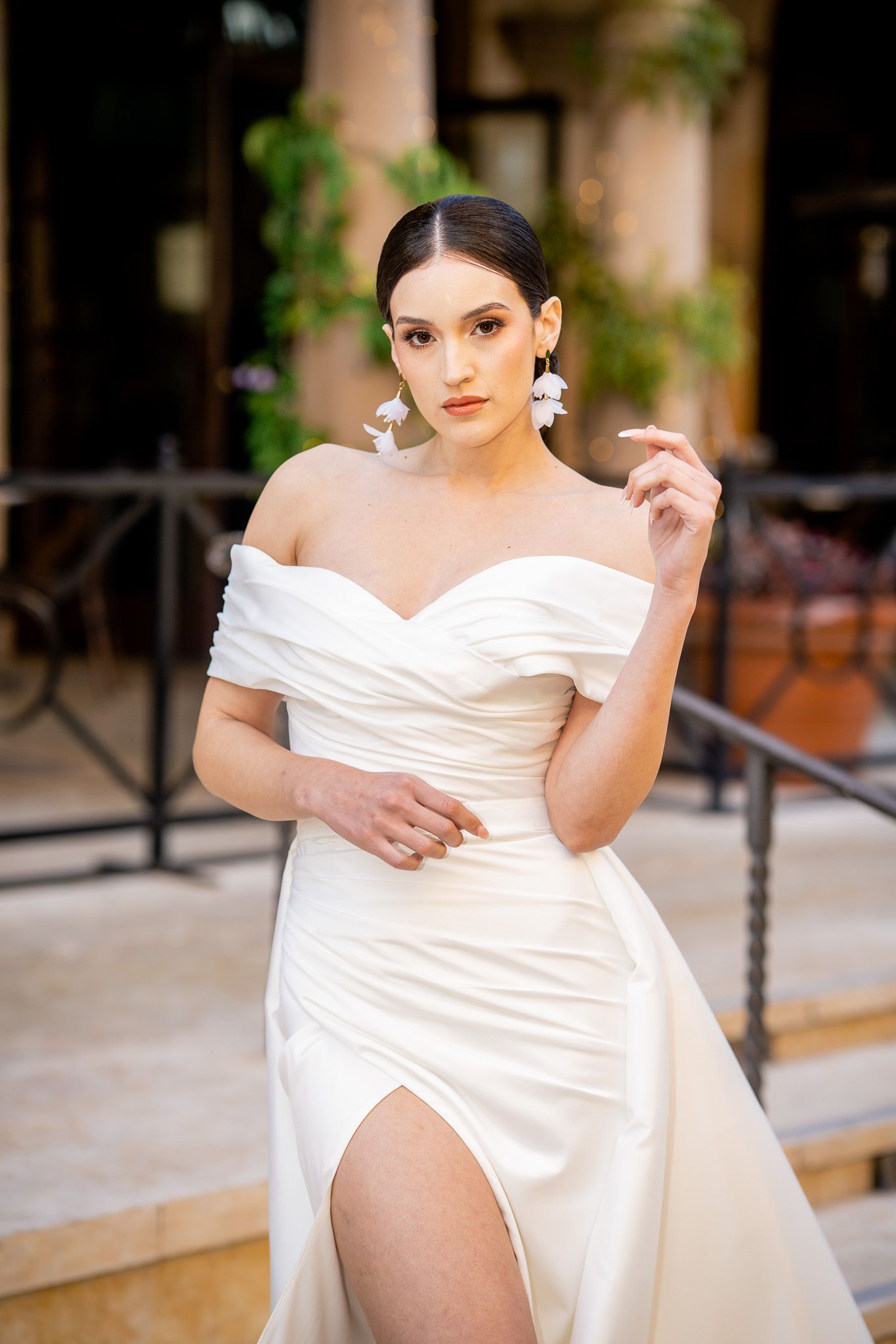 Elegant off-shoulder white wedding gown with thigh-high slit and sweetheart neckline in outdoor setting with stone columns.