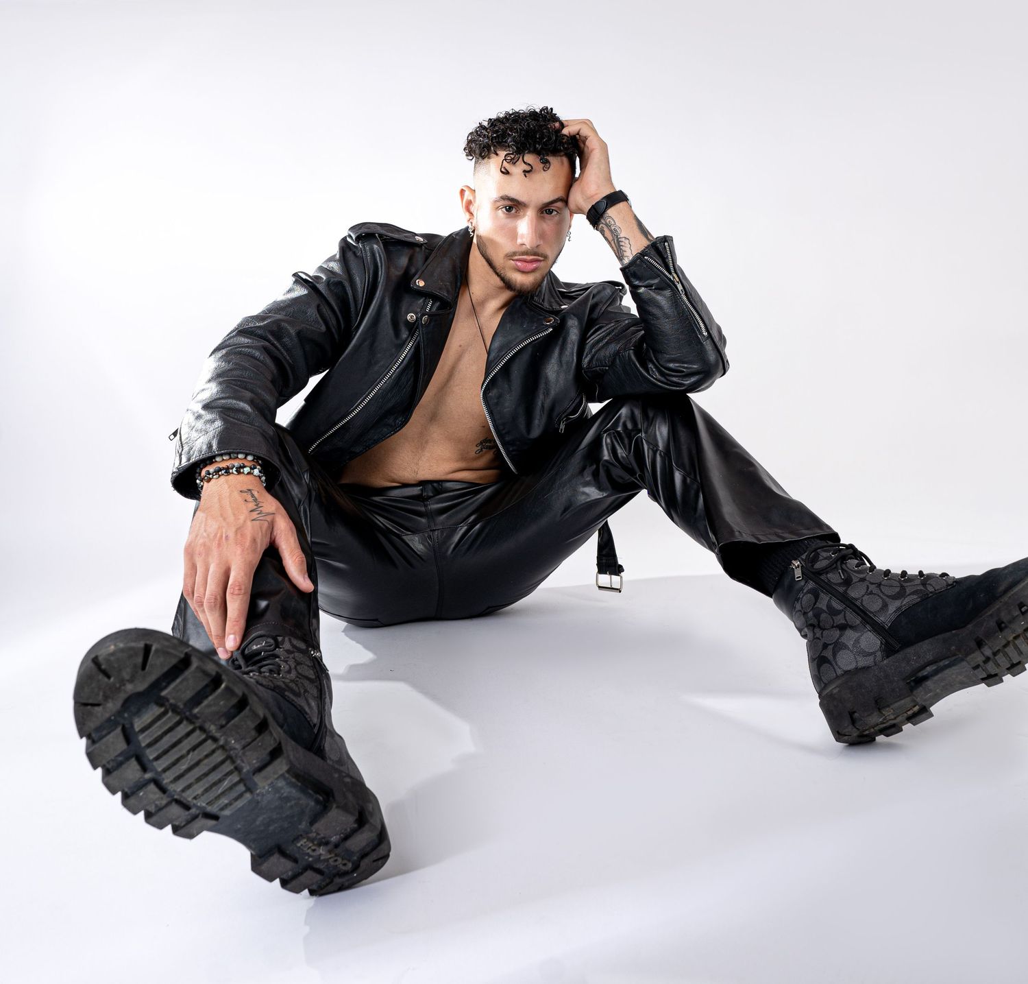 A model in a black leather jacket and combat boots poses dramatically on a white studio backdrop.