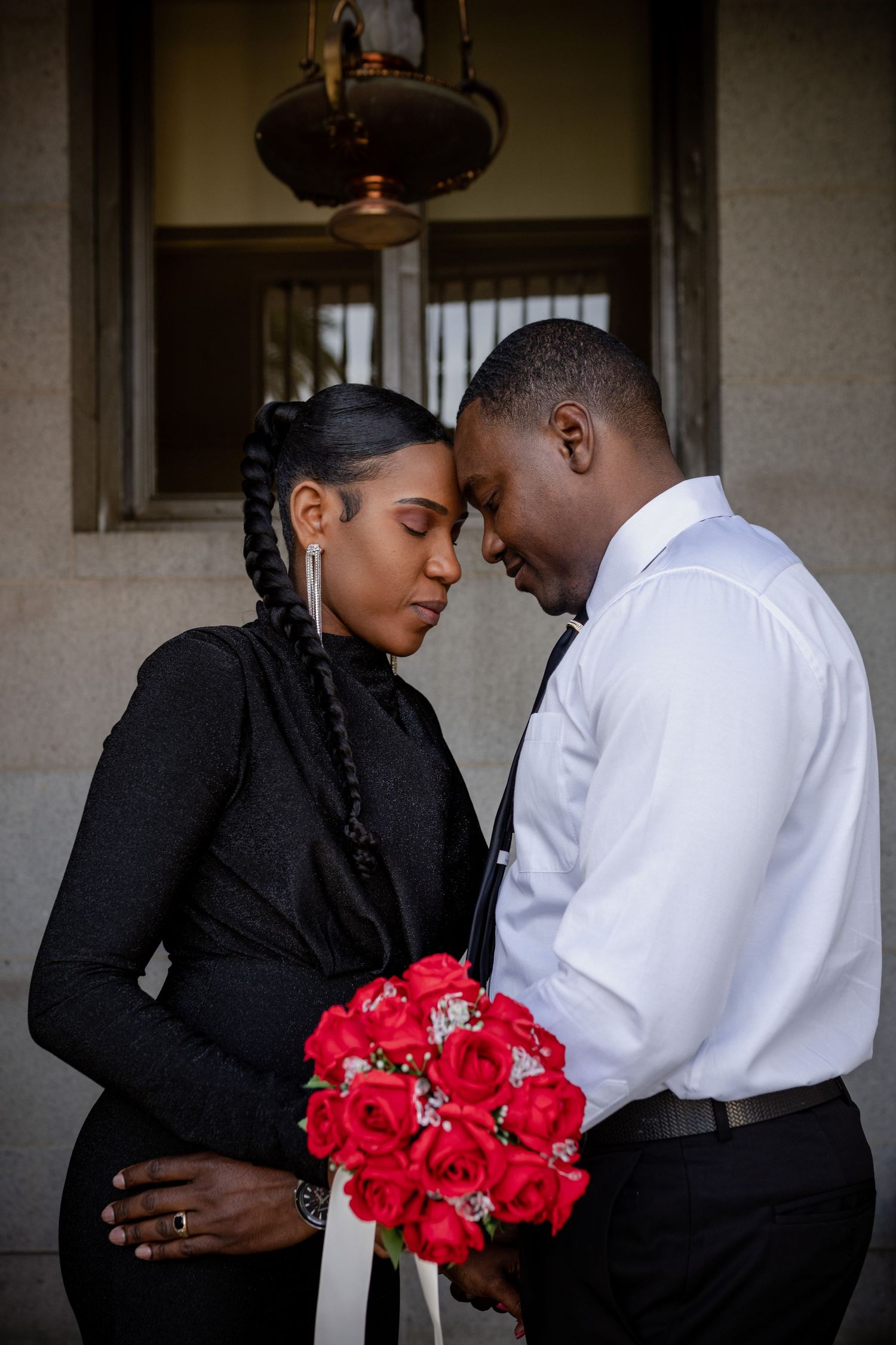 A romantic couple shares an intimate moment outdoors while holding a vibrant bouquet of red roses.