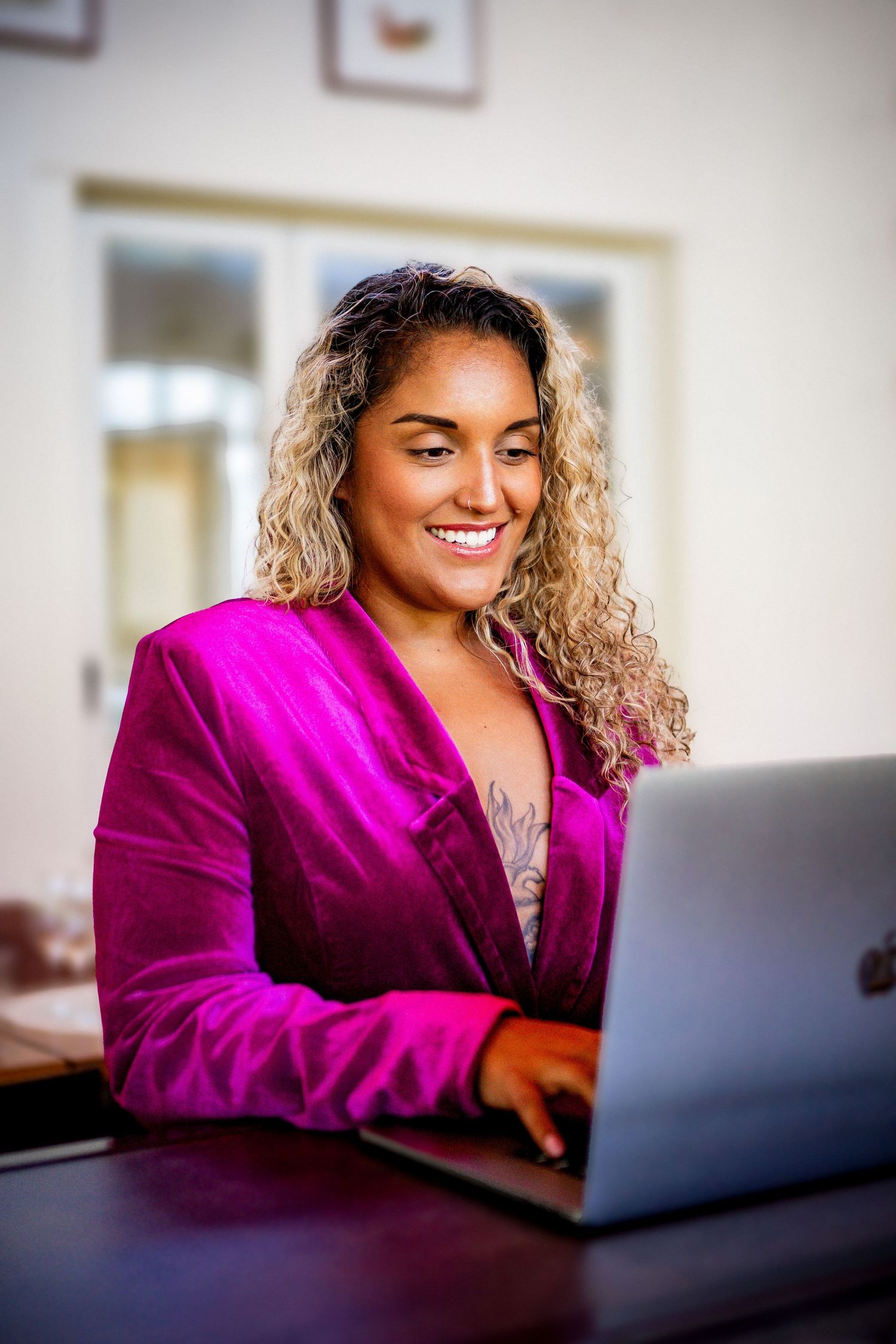 Professional working on laptop computer while wearing vibrant magenta blazer and smiling warmly in home office setting.