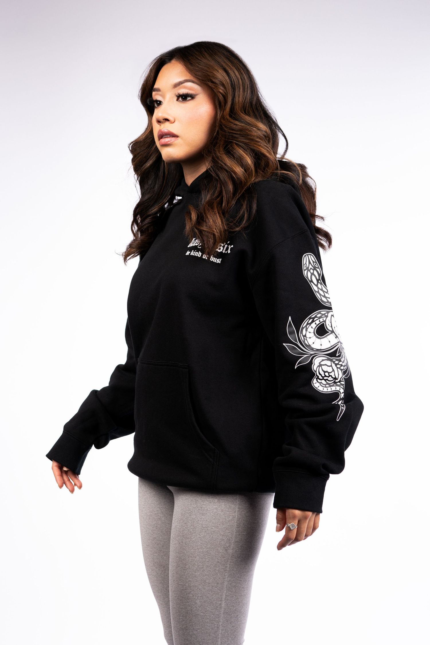 Person in black hoodie with white sleeve designs poses against light background while wearing gray leggings.