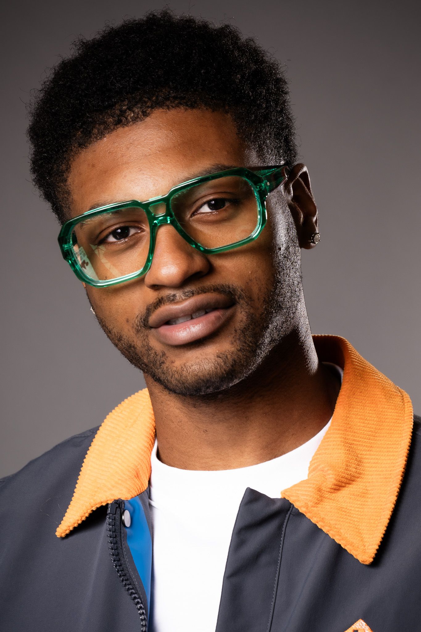 Stylish portrait of a person in green glasses and an orange collared jacket against a dark gray background.
