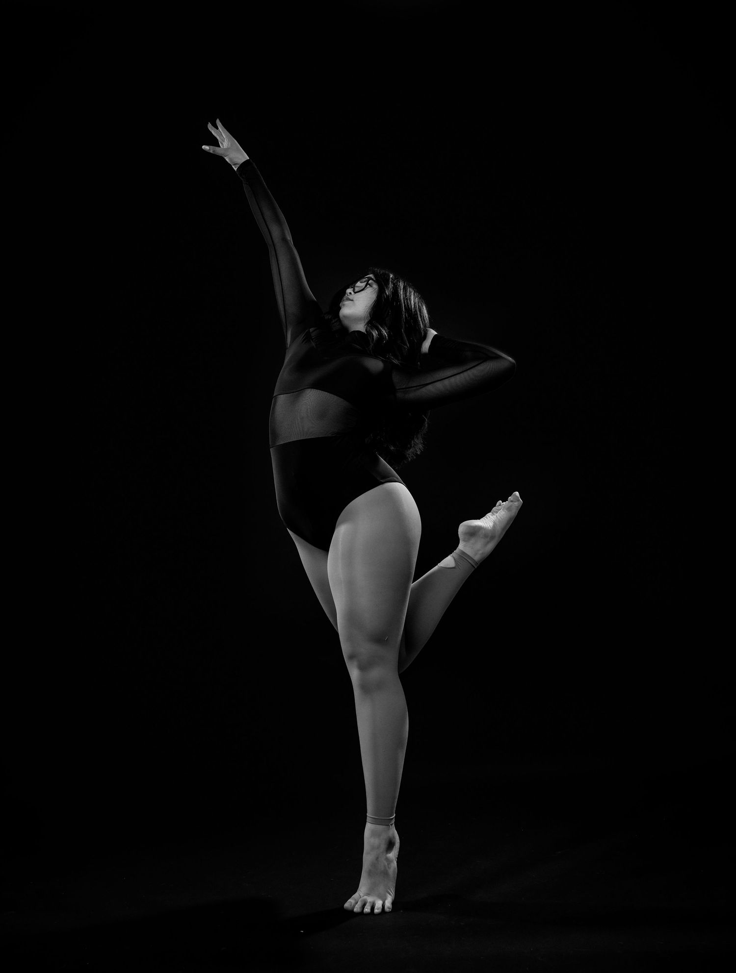 A dancer in a black leotard performs a dynamic pose against a dark background in black and white photography.