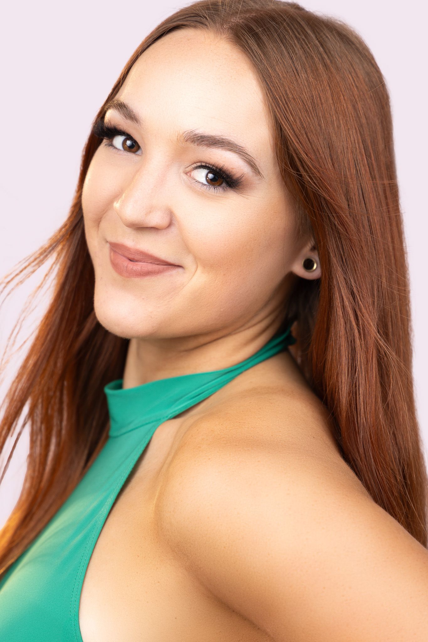 Portrait of someone with long brown hair and subtle makeup wearing an emerald green halter top against a pink background.