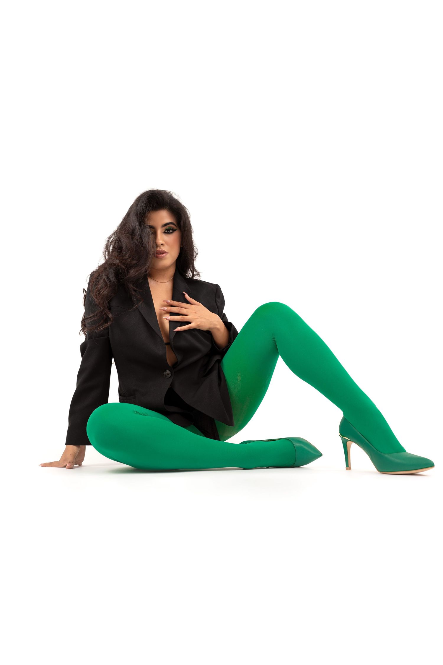 Woman in black blazer and vibrant green tights poses elegantly on floor in stylish outfit.