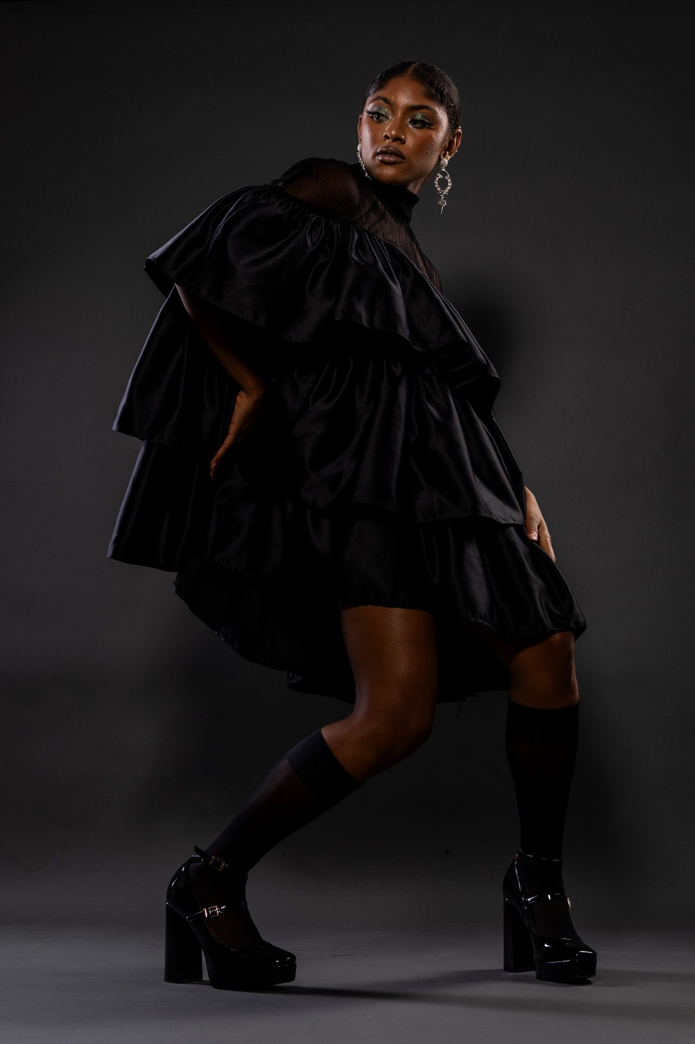 Dramatic fashion portrait showing black ruffled dress with platform boots against dark background in studio lighting.