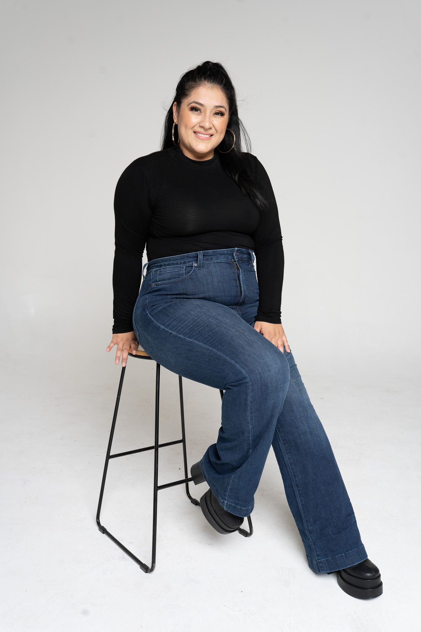 Person in black long sleeve top and wide leg jeans poses on modern black barstool against light gray background.