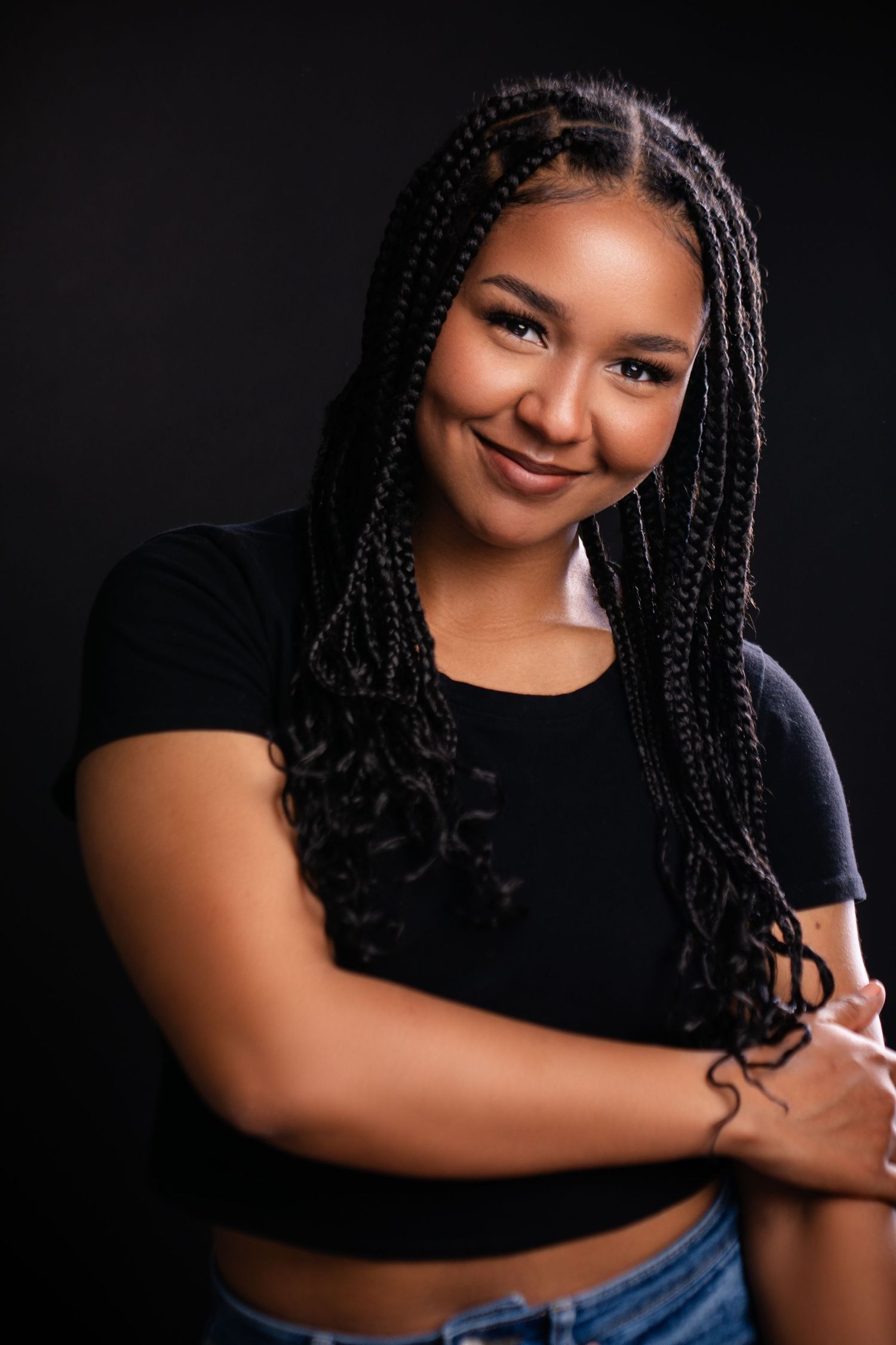 Studio portrait with black top and jeans featuring long braided hair with curled ends against dark background.