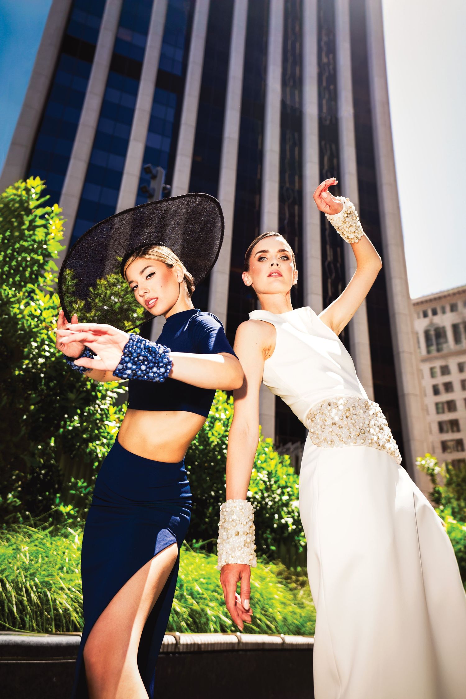 Two fashionable models pose dramatically in contrasting outfits against an urban backdrop with modern buildings and greenery.
