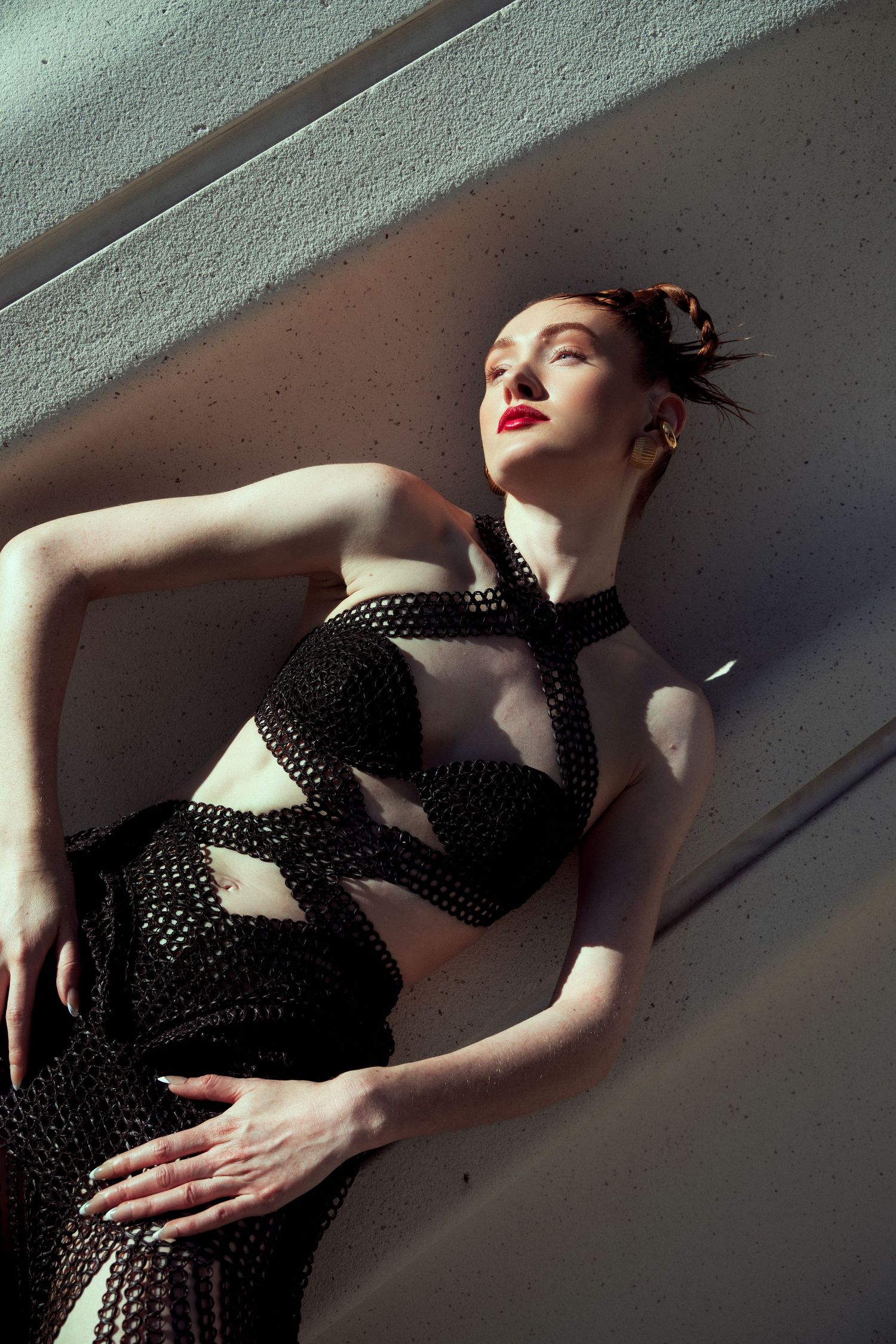 Dramatic fashion photograph of a figure in a black cutout dress with red lipstick posing against an angled wall in shadow.