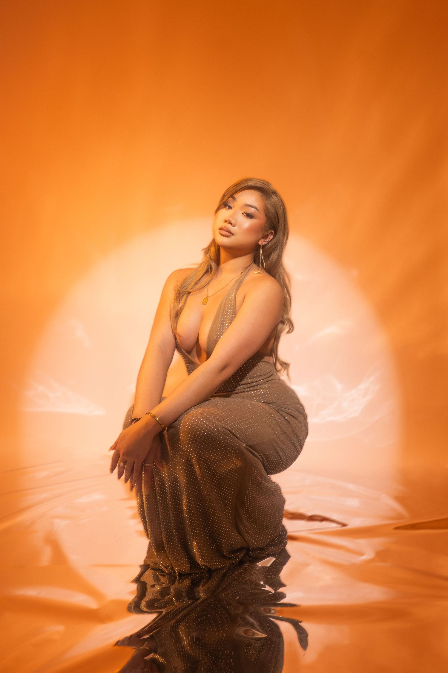 Fashion portrait in warm orange tones with figure in flowing brown dress posing against sunset-like backdrop.