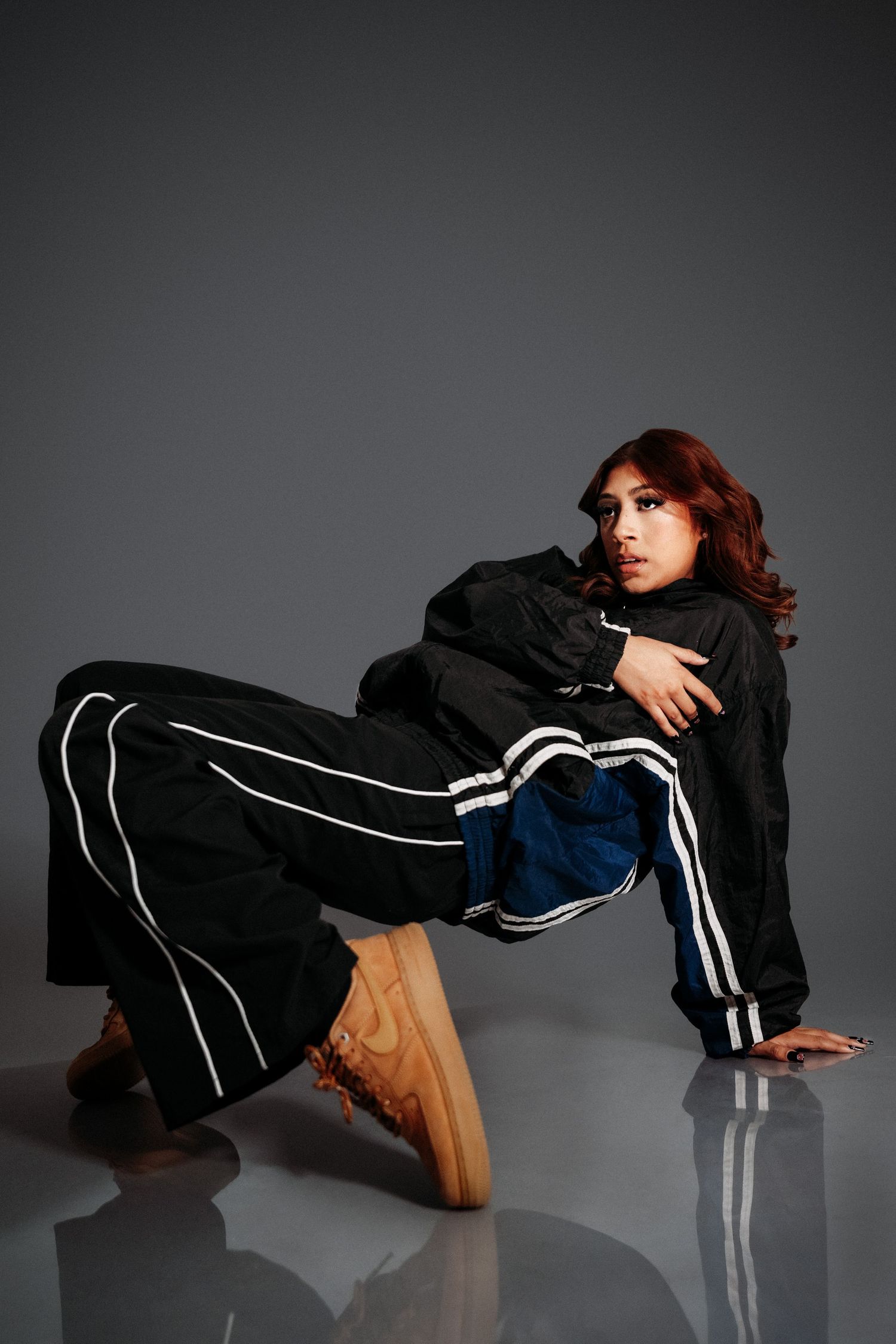 A person in black tracksuit with white stripes strikes a dynamic pose while wearing tan sneakers on reflective surface.