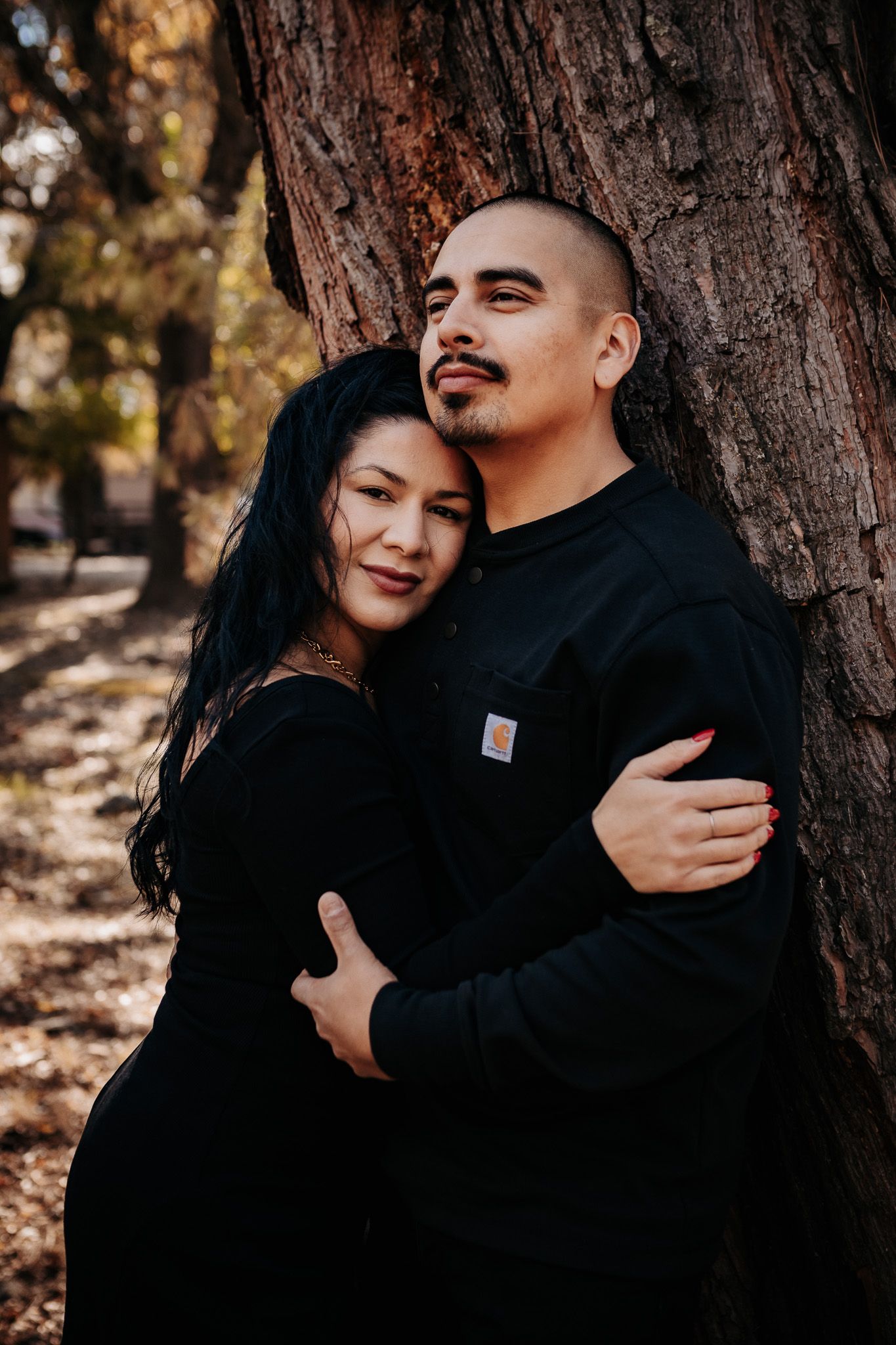 A couple embraces against a large tree trunk on an autumn day, both wearing matching black sweaters.