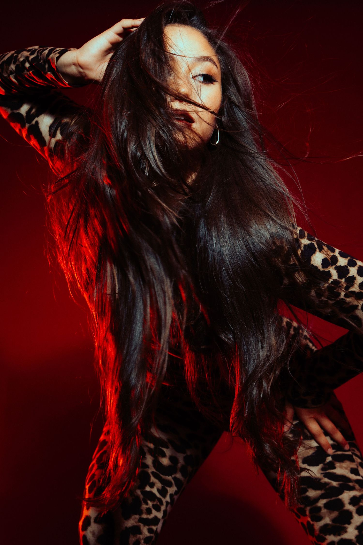 Dynamic fashion portrait with flowing dark hair against red backdrop wearing animal print outfit in dramatic lighting.