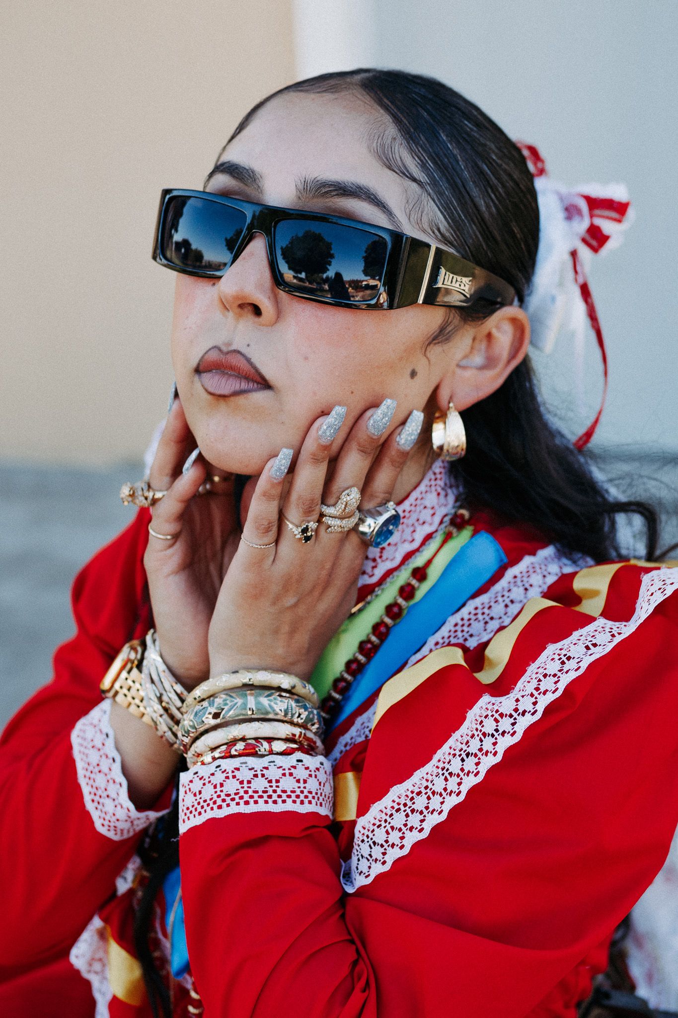 A fashion portrait featuring stylish rectangular sunglasses, multiple rings, and a vibrant red traditional outfit with lace details.
