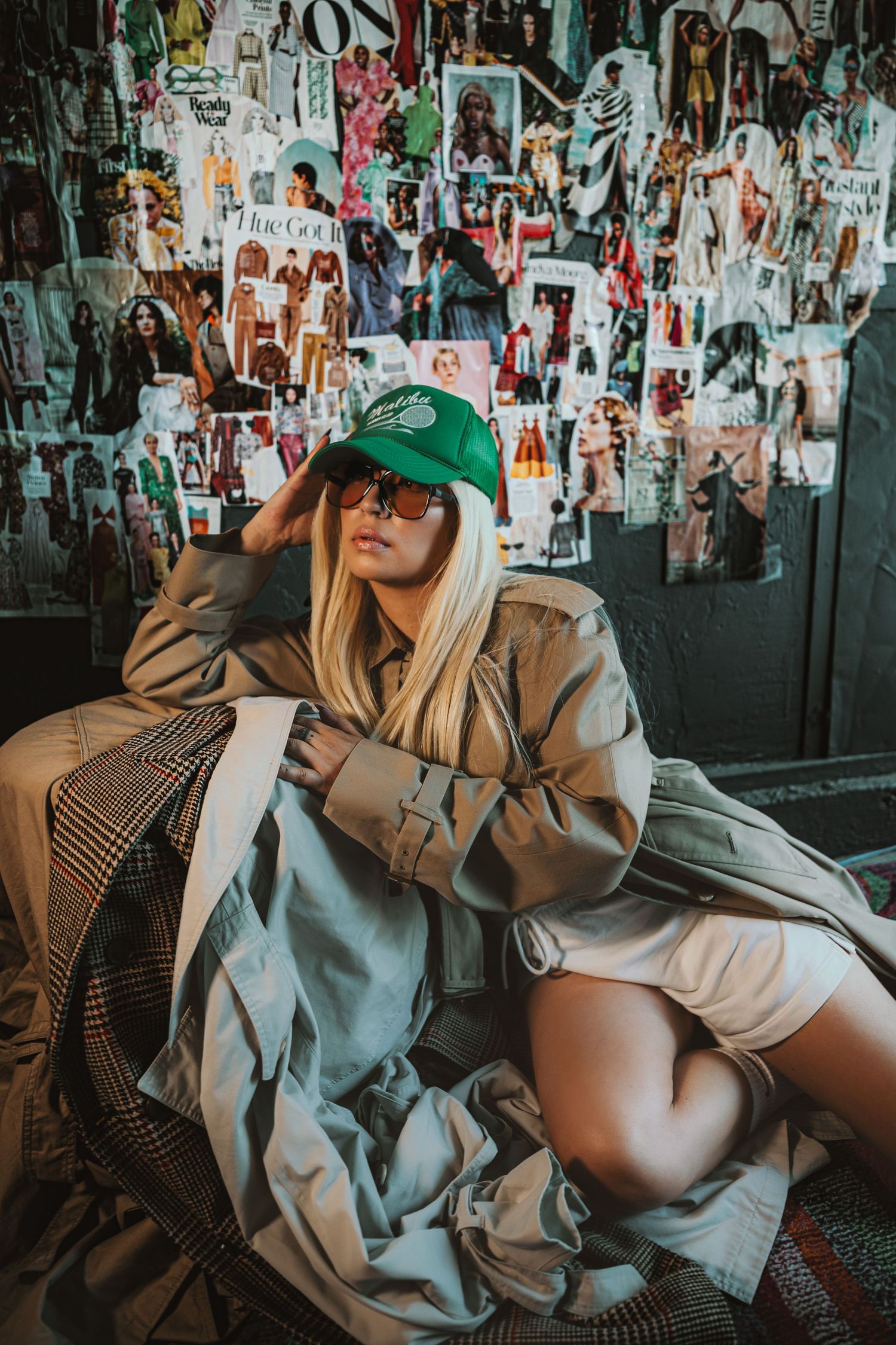 Street style fashion photo with person in oversized beige trench coat, green baseball cap and biker shorts against graffiti wall.