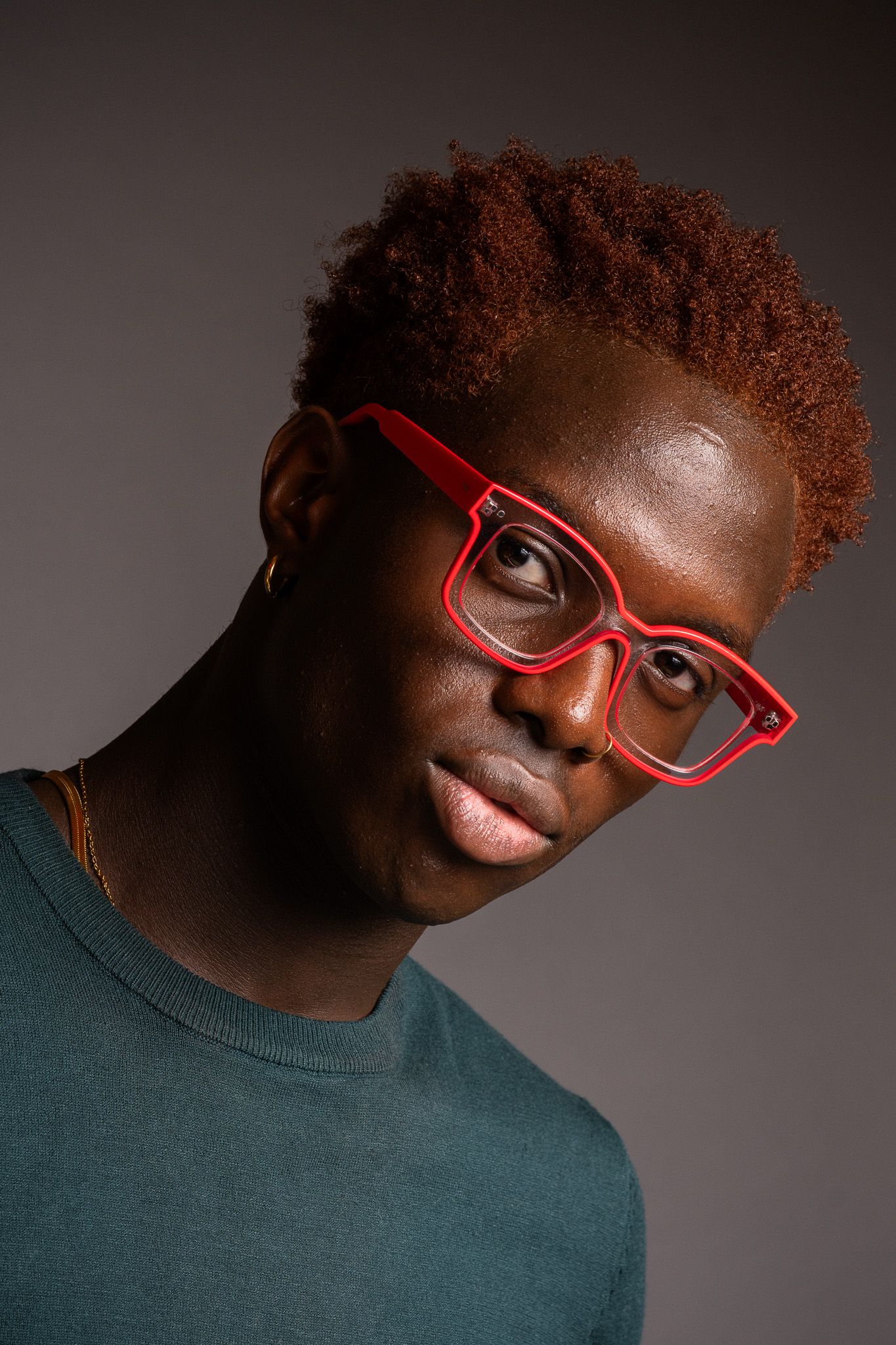 Artistic portrait of a person with red-tinted hair wearing bright red glasses and a dark green top.