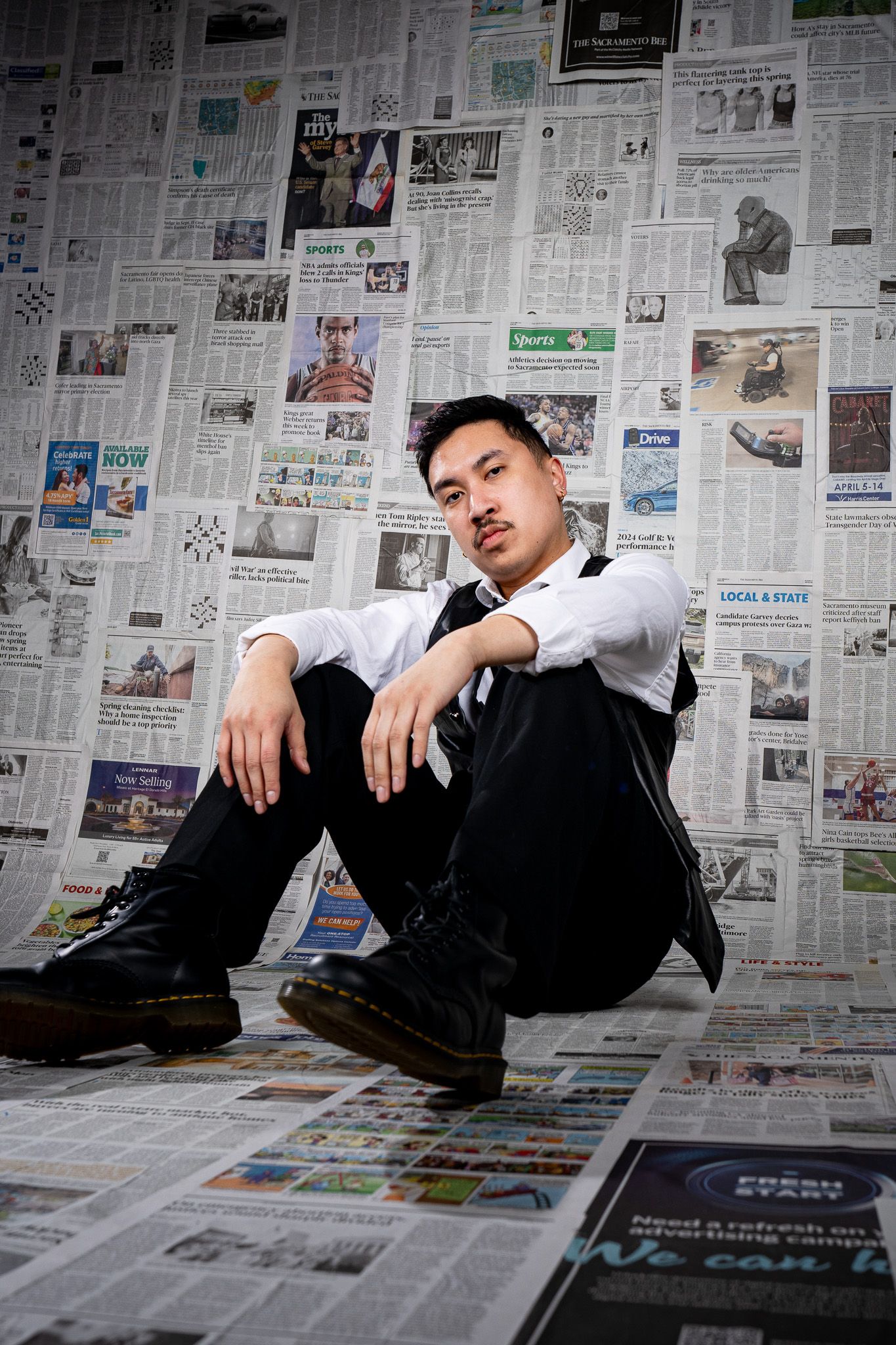 An artistic portrait of someone sitting against a wall covered in newspaper clippings wearing a white shirt and black pants.