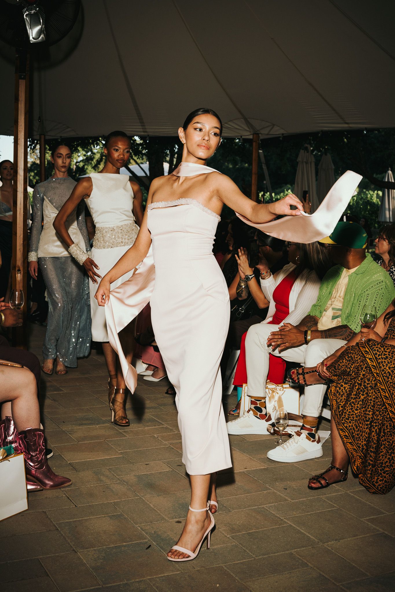Models walk the runway in sleek white dresses and high heels during an outdoor fashion show under a tent.