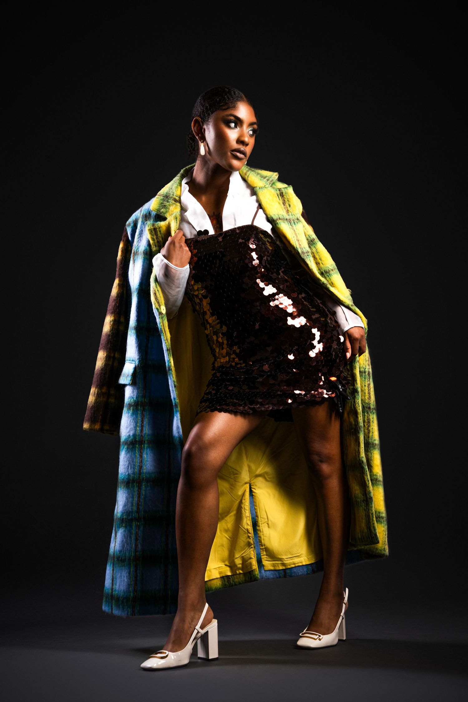 Fashion model in sequin dress and bold yellow plaid coat poses dramatically against dark background with white heels.