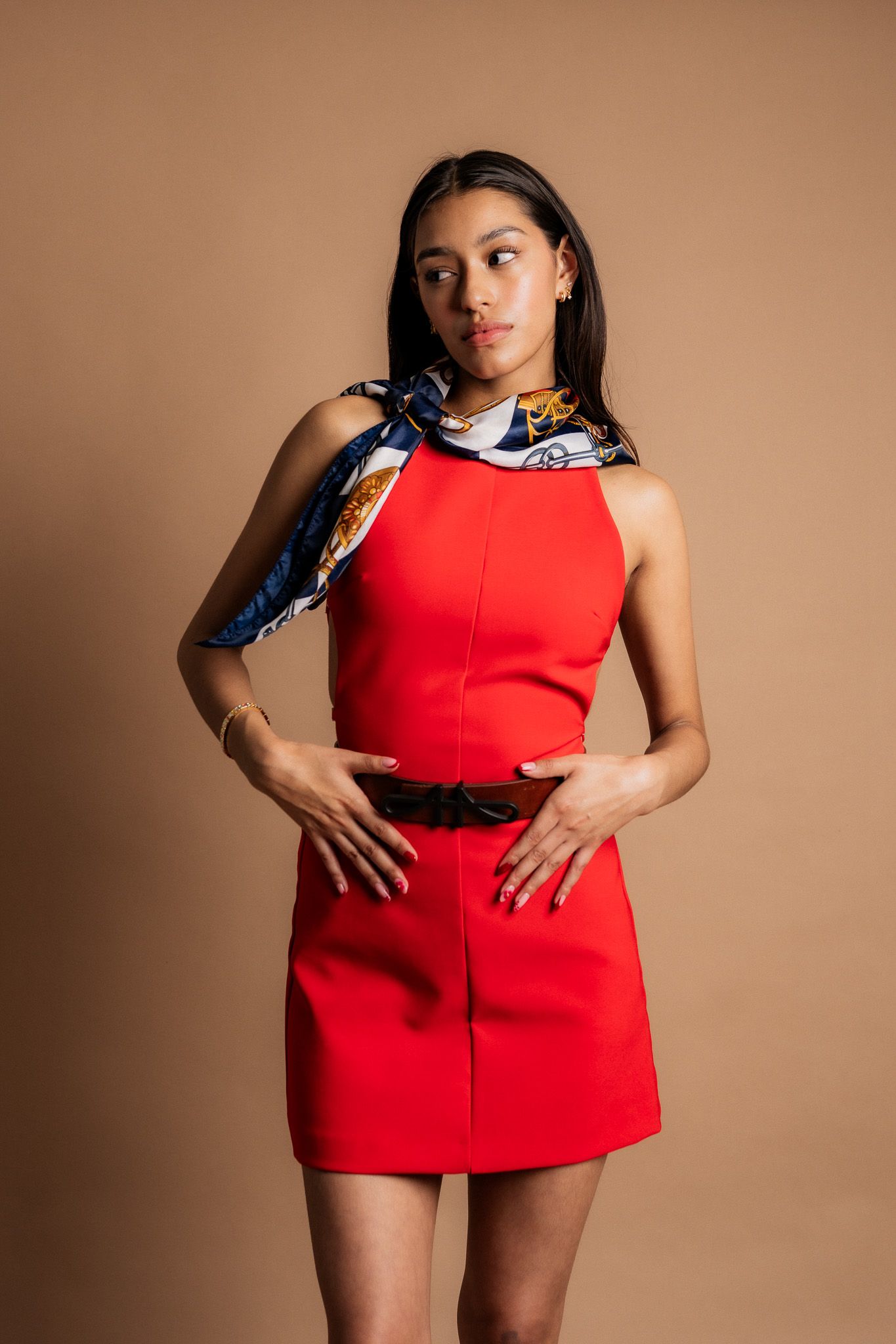 Person in a vibrant red sleeveless dress poses against brown background with blue patterned neck scarf.