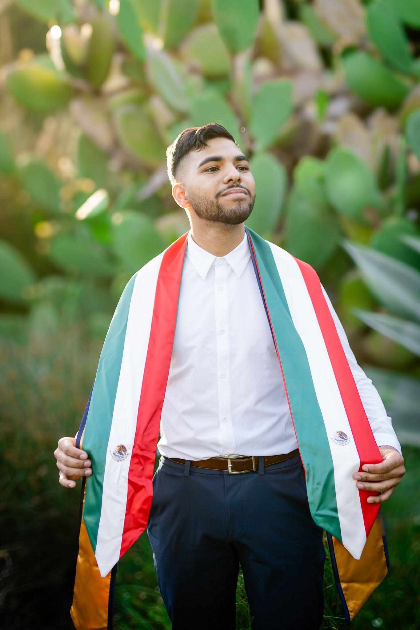 Someone wearing a white dress shirt and navy pants drapes a Mexican flag around their shoulders in an outdoor setting.
