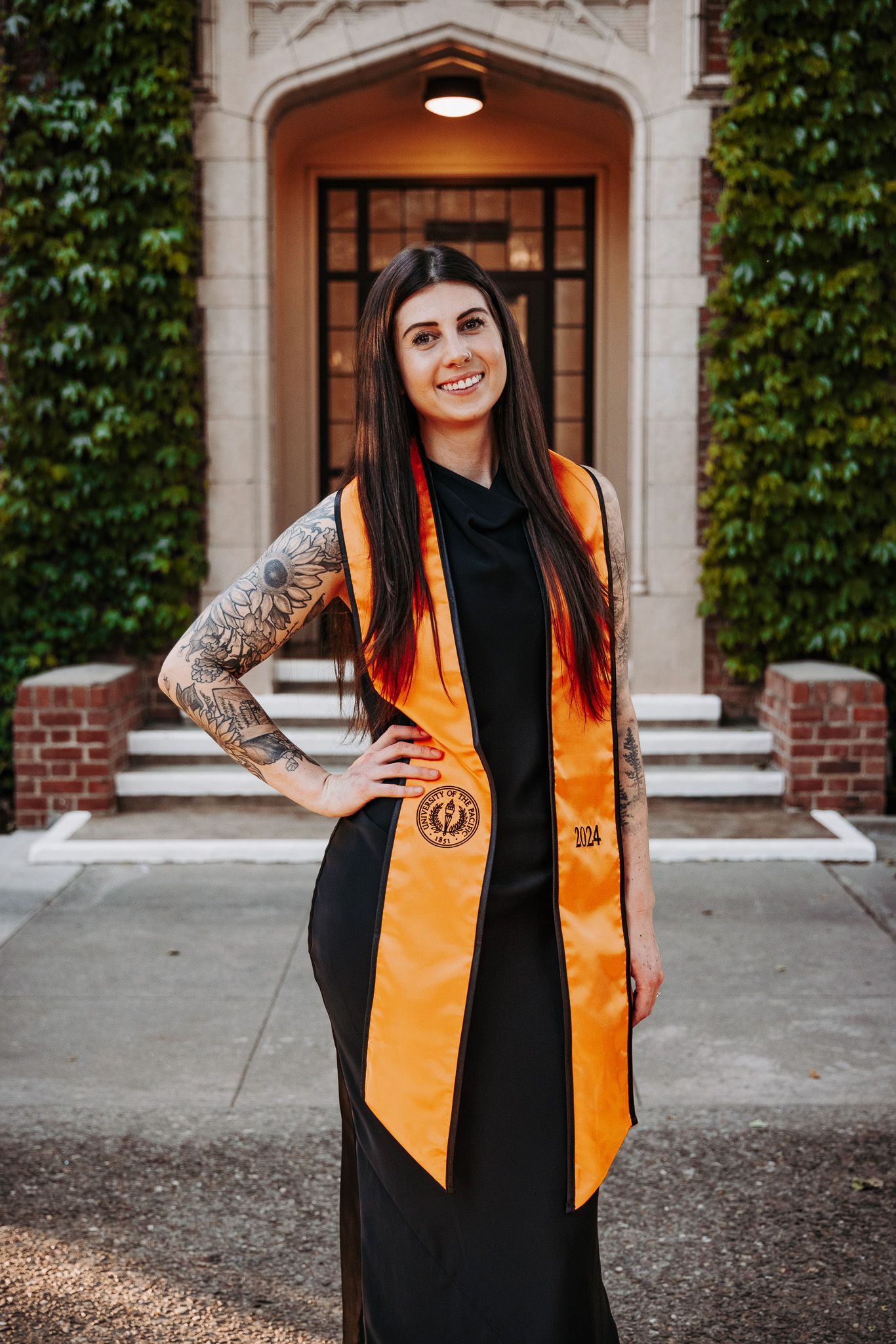 Graduate wearing black graduation gown and orange honors stole stands in front of ivy-covered doorway on campus.