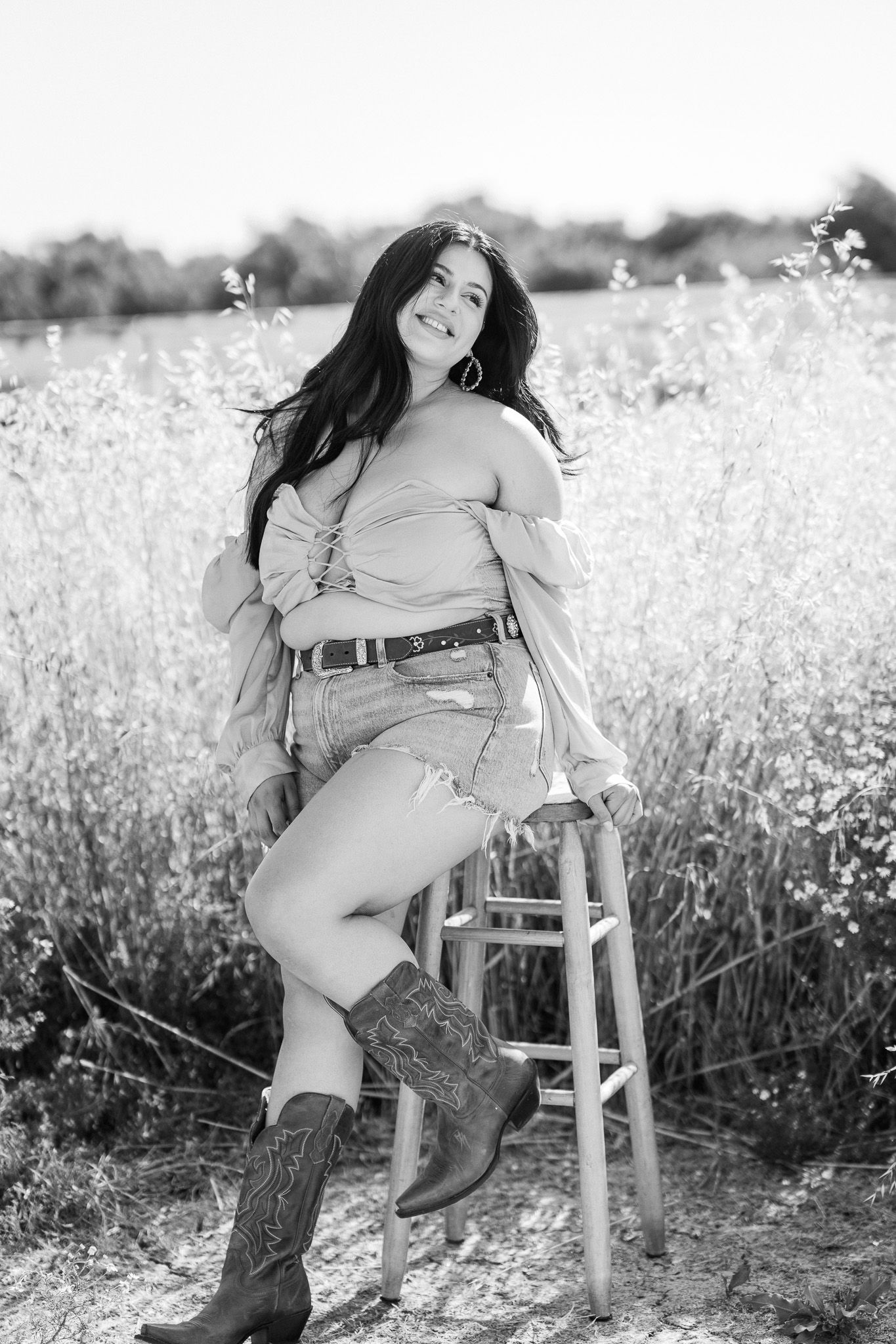 Black and white portrait of person in cowboy boots and off-shoulder top posing on wooden stool in wildflower field.
