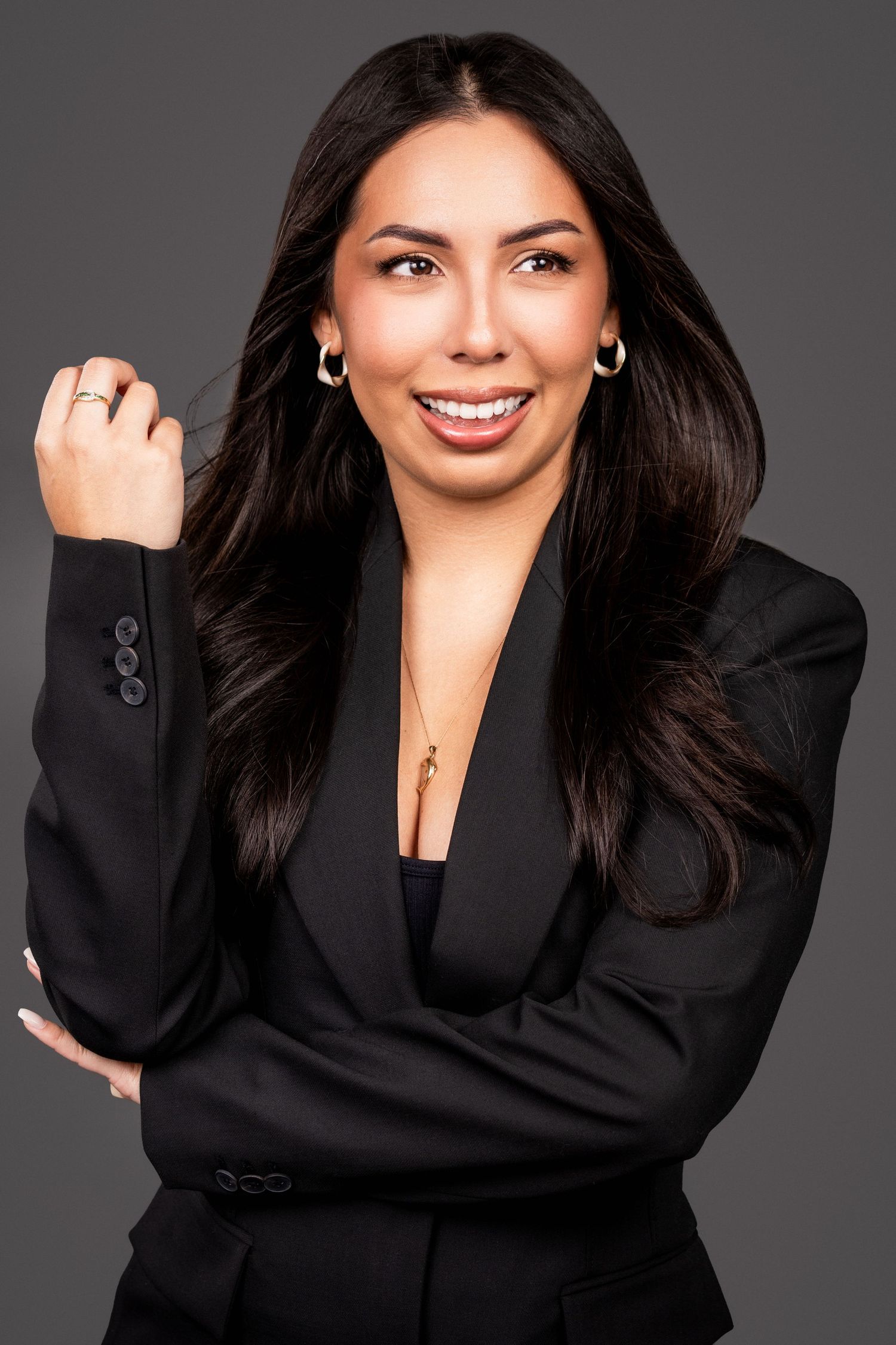 Professional business portrait with dark hair wearing a black blazer and smiling warmly against a gray background.