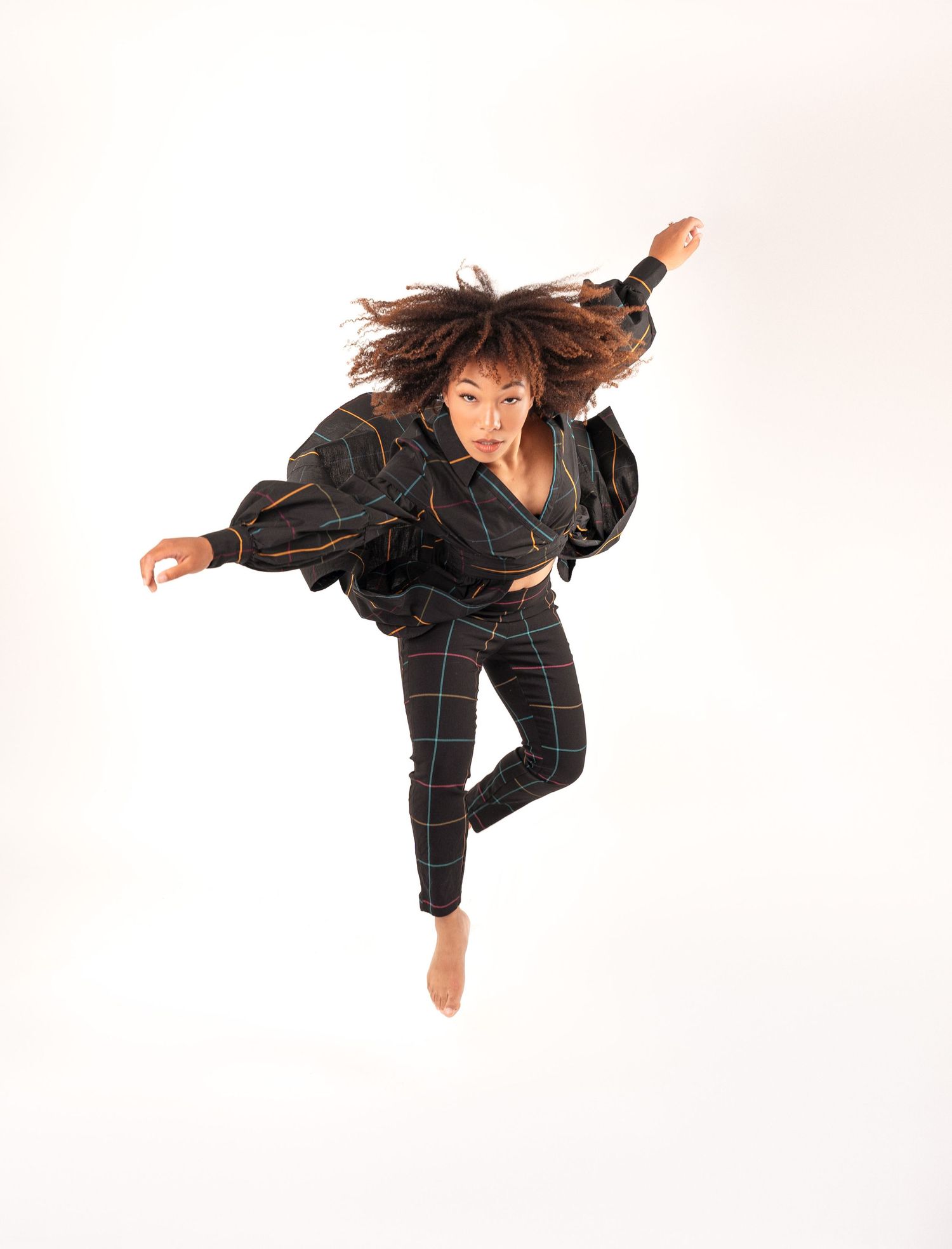 Contemporary dancer in black windowpane outfit leaps through the air against white background, hair flowing dynamically.