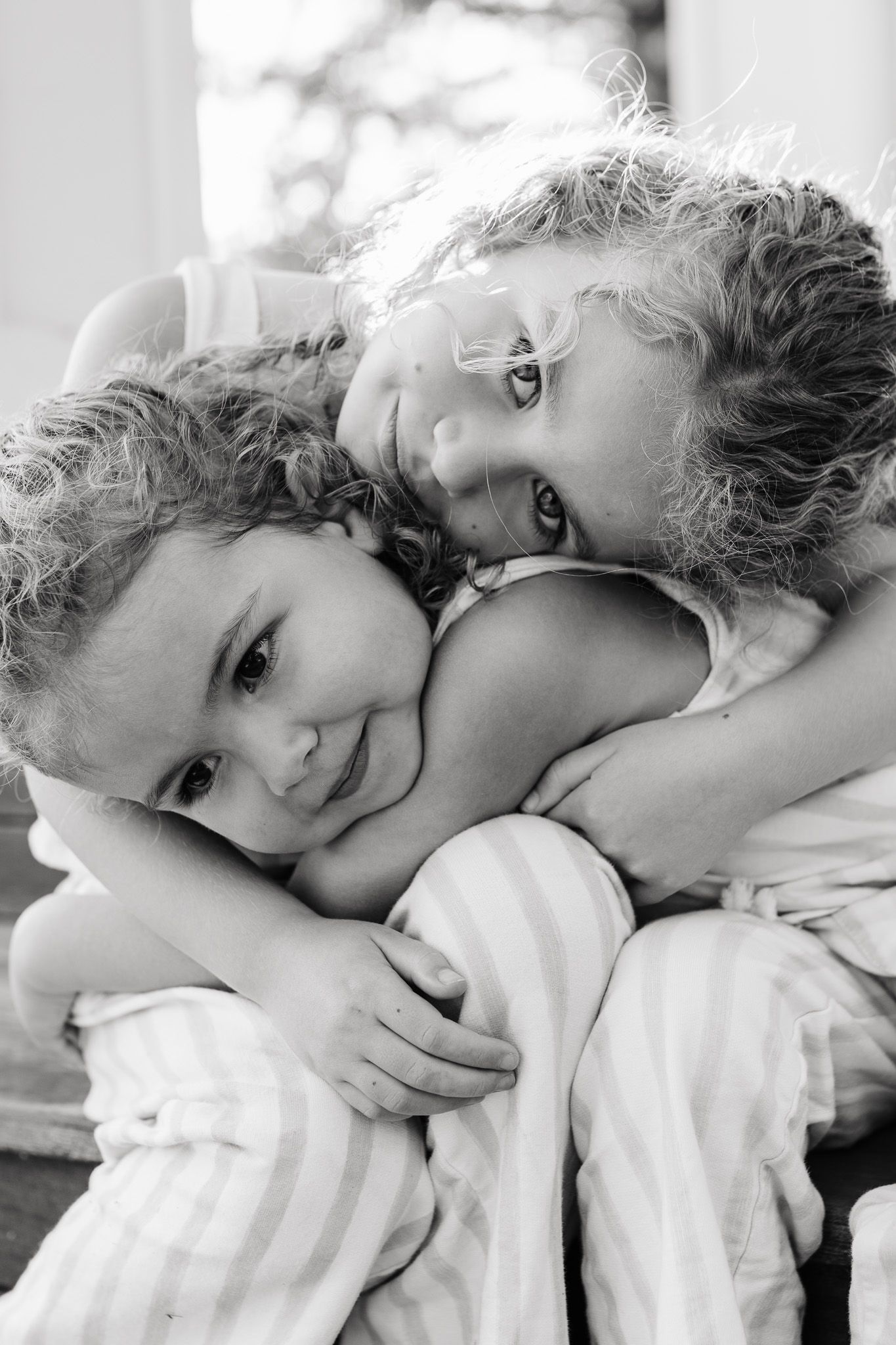 Black and white intimate portrait of two young children embracing while resting on soft bedding.