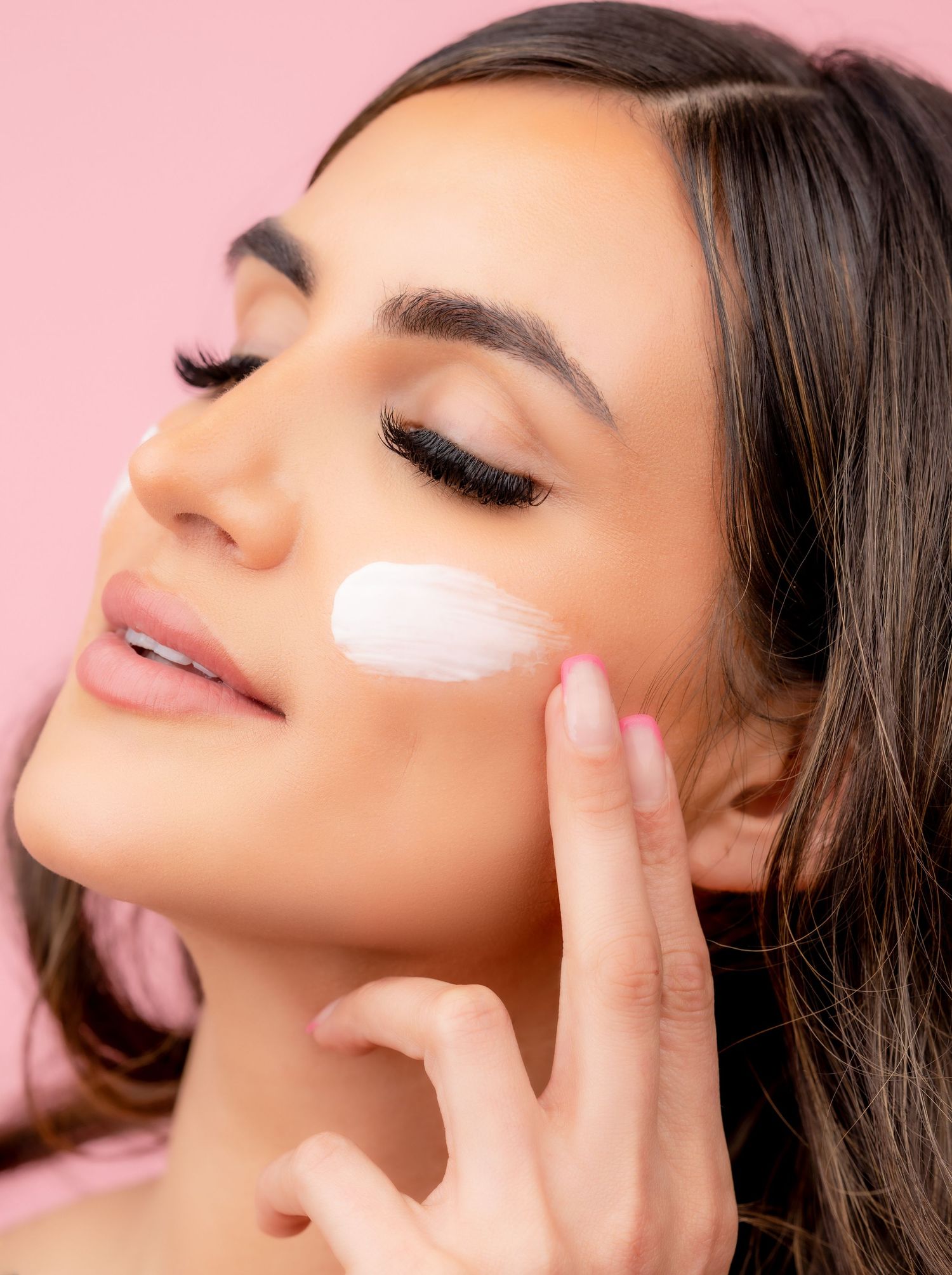 Woman with flawless makeup applying moisturizer cream on her face against pink background.