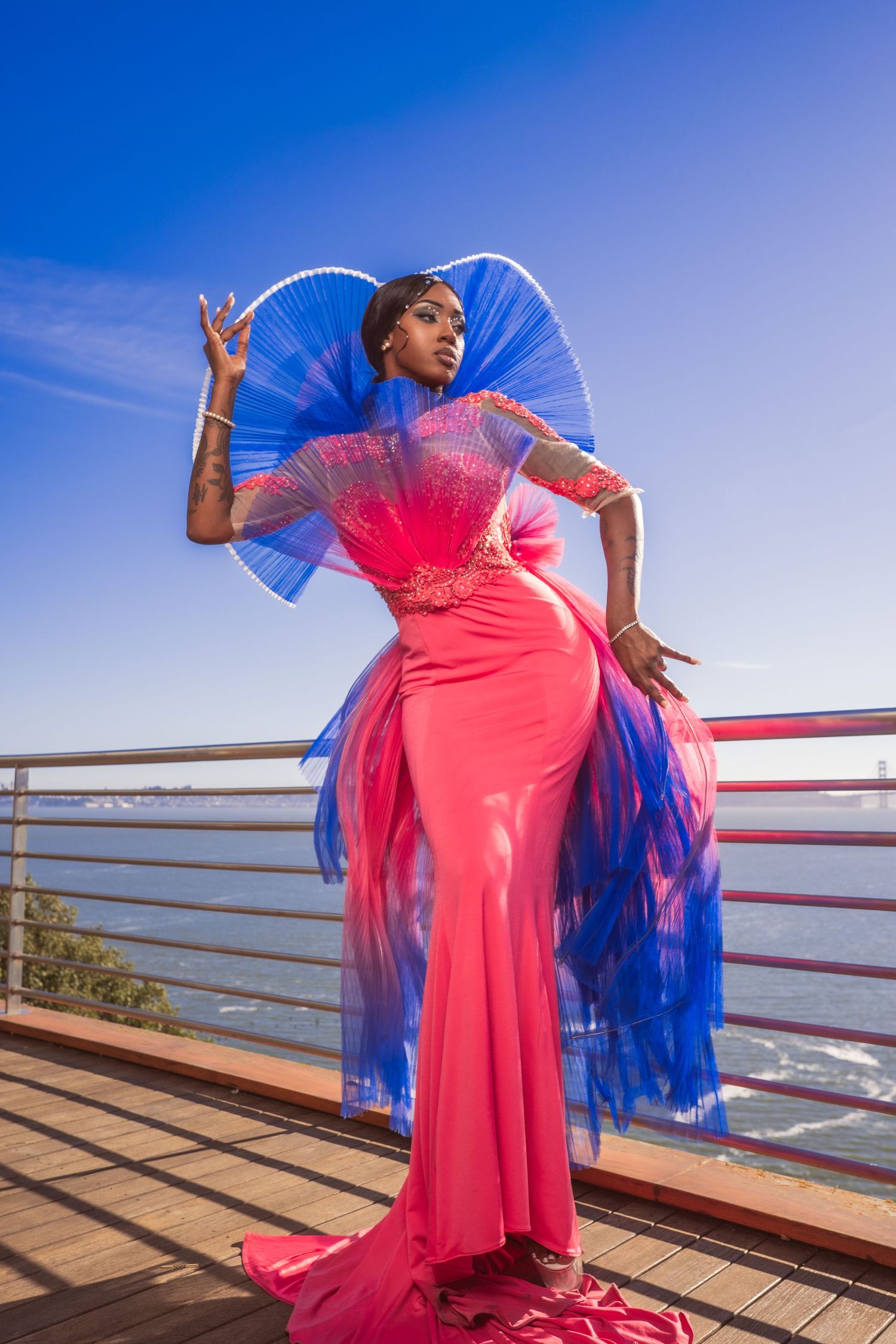 A person in a stunning pink gown with blue tulle accents poses dramatically on an outdoor deck against a bright blue sky.