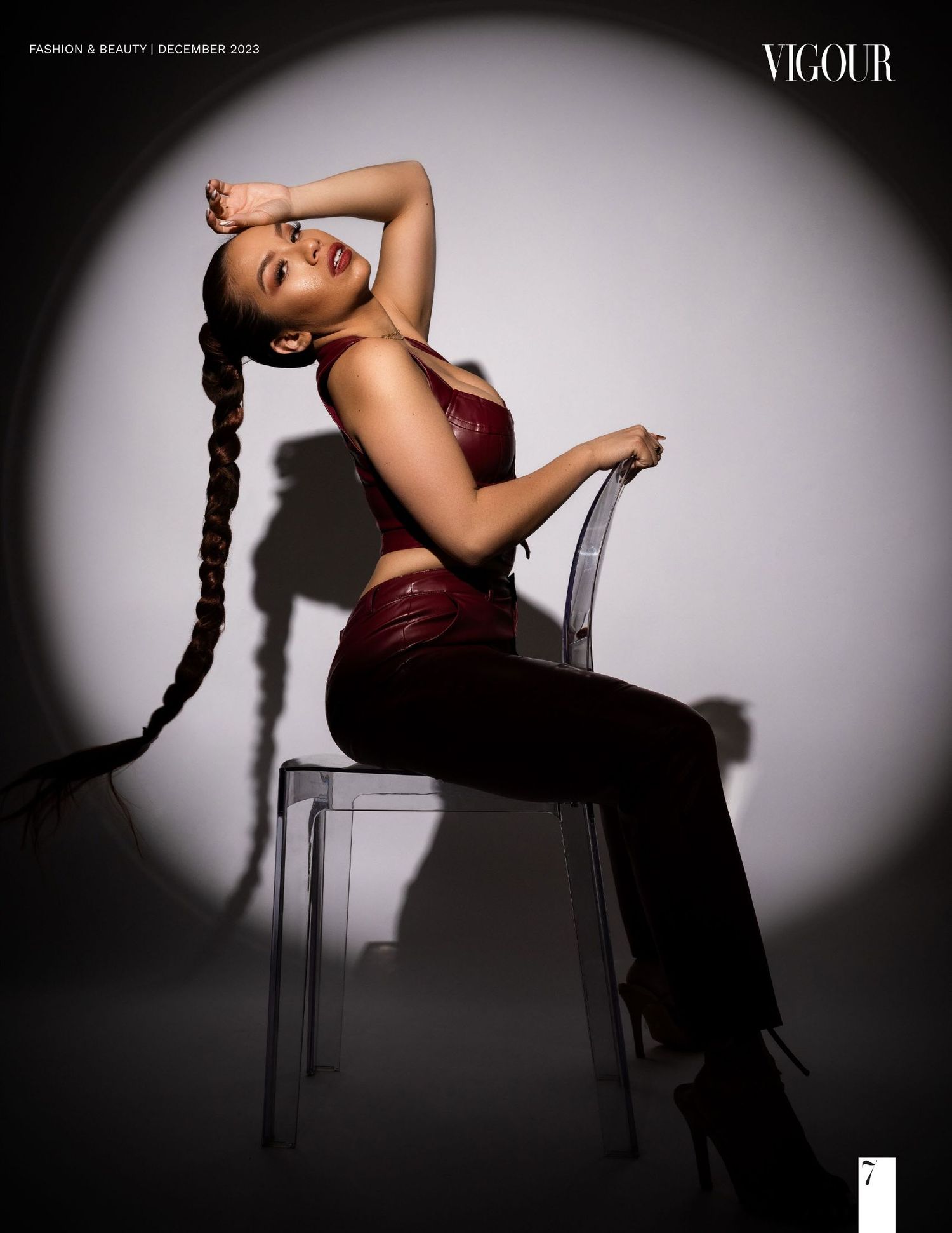 Fashion portrait in dramatic lighting showing model in burgundy outfit posing on chair with long braided hair.