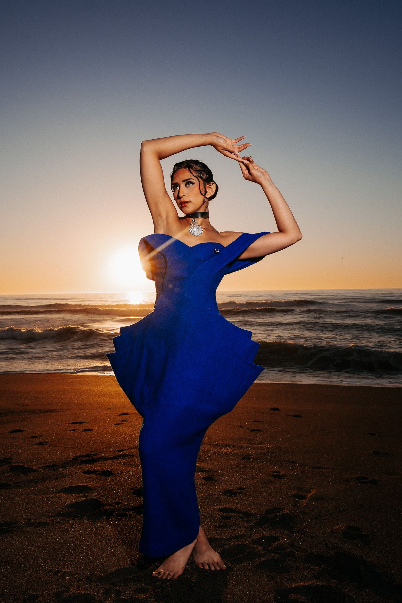 Woman in flowing blue dress poses gracefully on beach at sunset with arms raised.