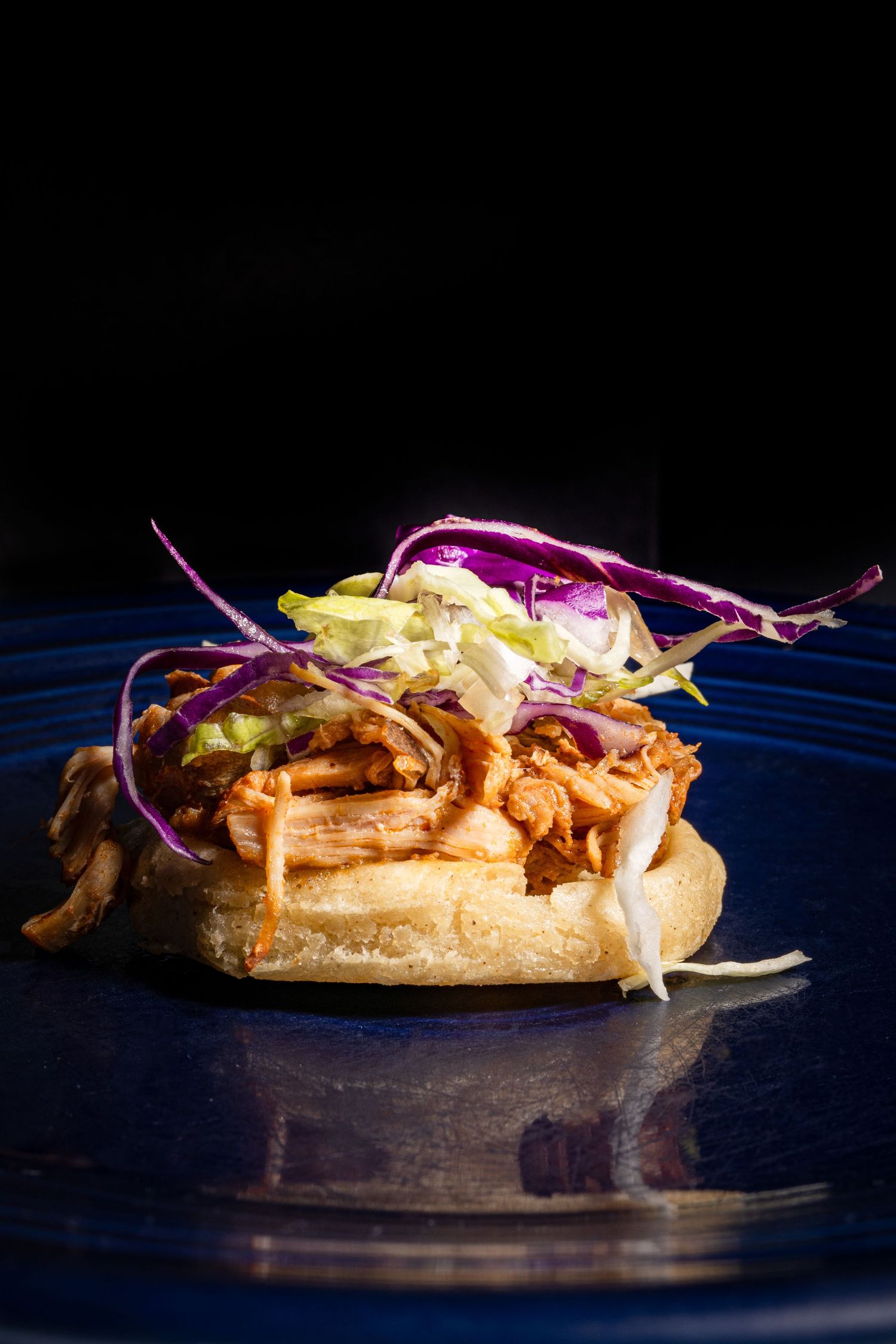 A gourmet pulled pork sandwich topped with colorful purple and yellow coleslaw on a blue plate against dark background.