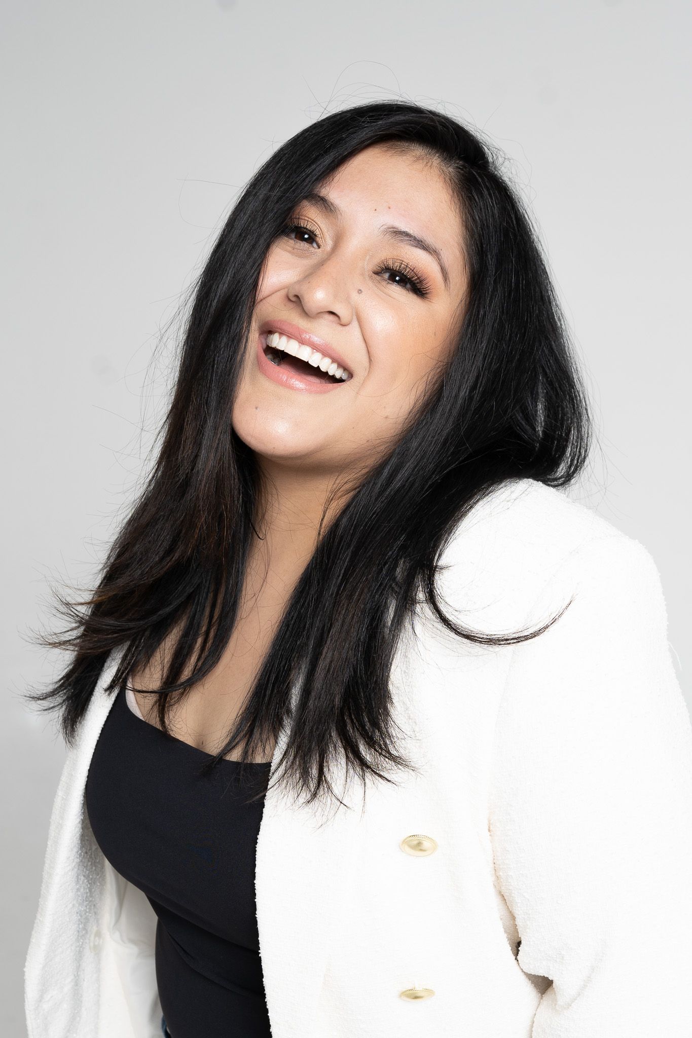 Portrait of someone with dark hair wearing a black top and white blazer laughing joyfully against light background.