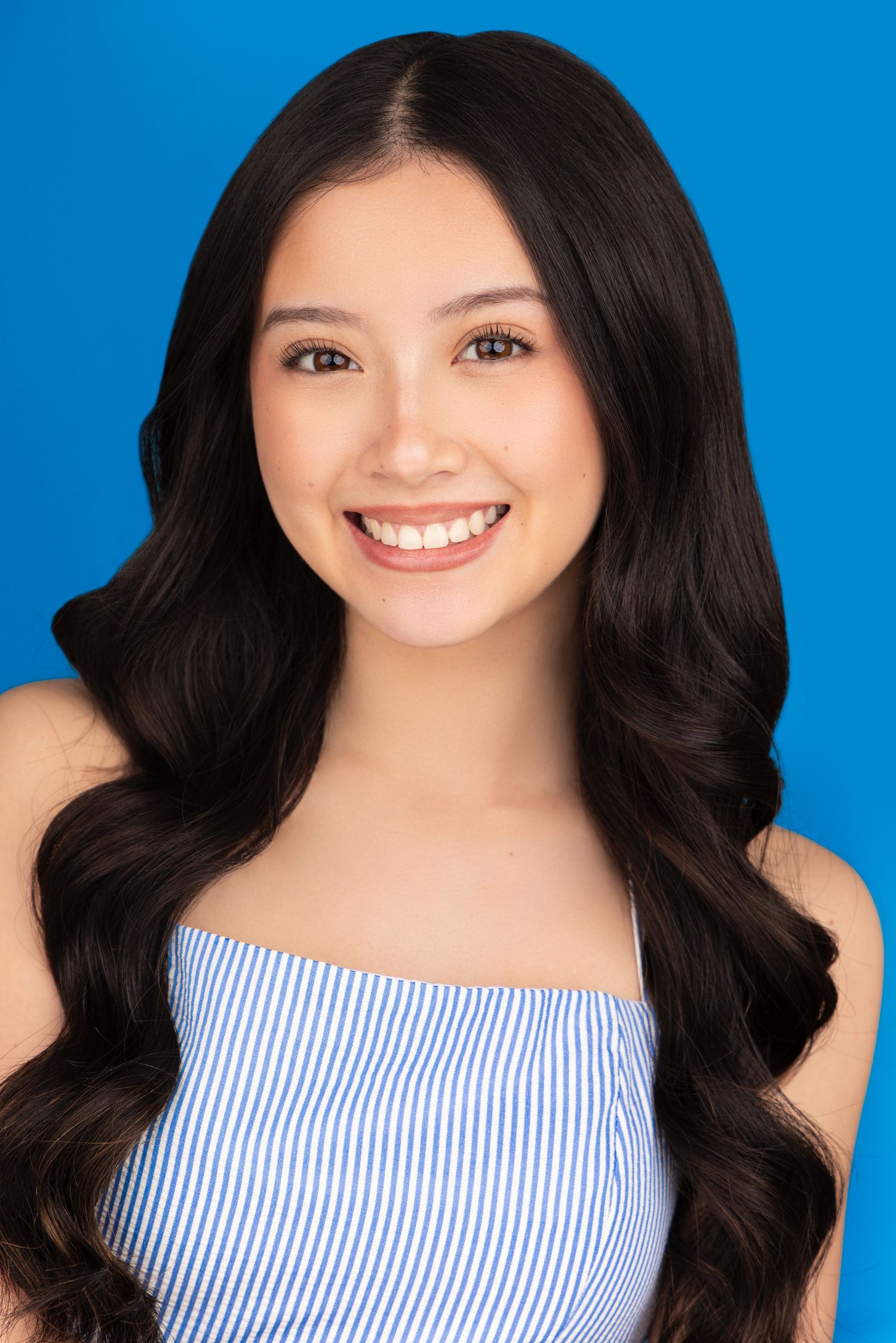 Professional portrait with long dark wavy hair wearing a striped off-shoulder top against a bright blue background.