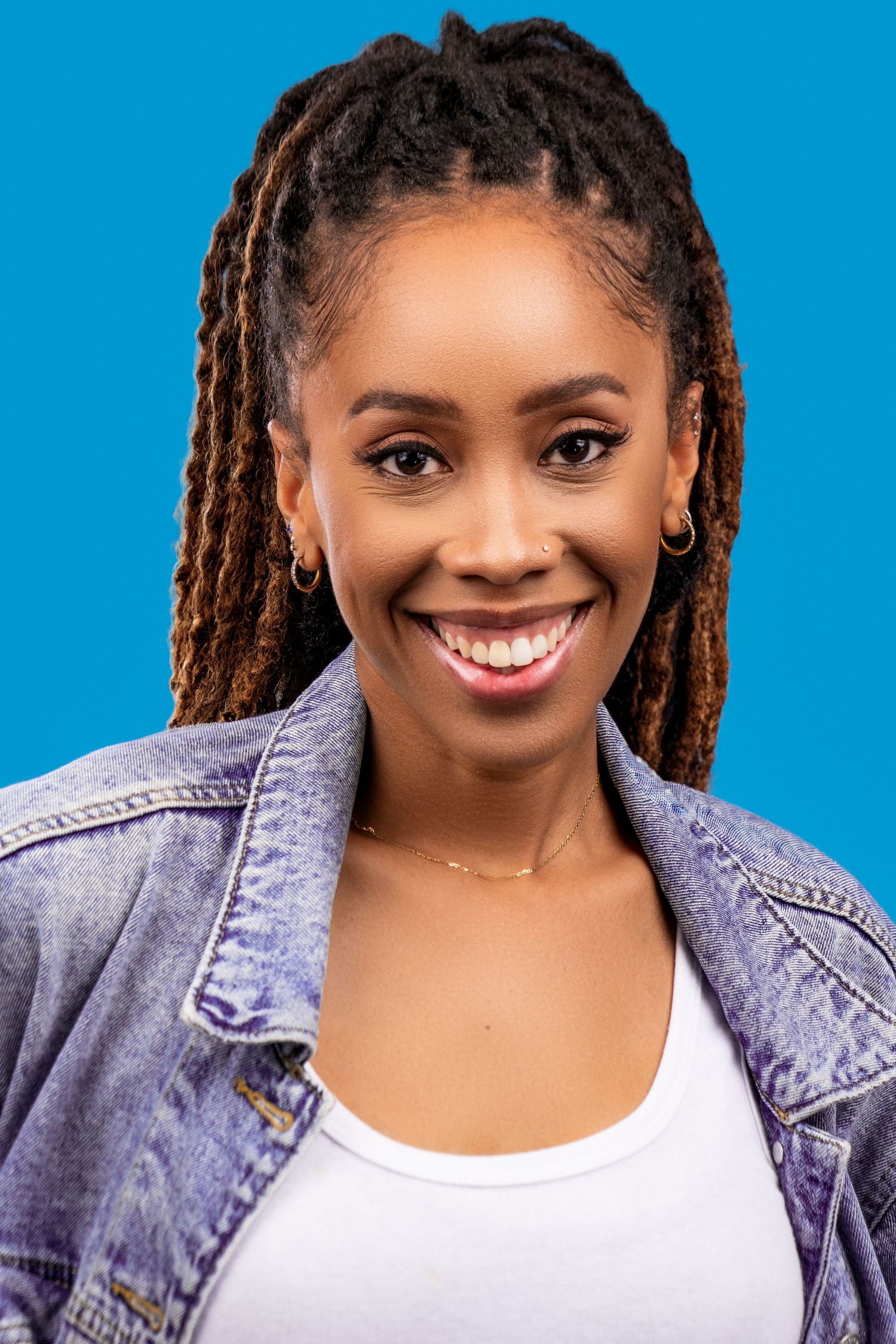 Portrait of someone with long locs hairstyle wearing a denim jacket and white top against a blue background, smiling warmly.
