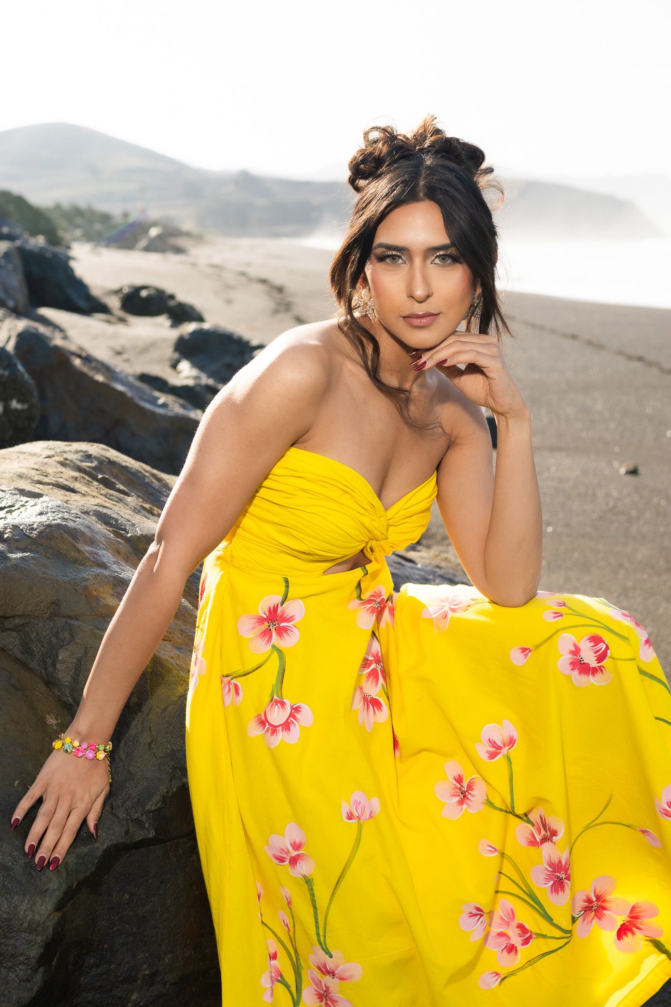 Yellow strapless dress with pink floral details photographed on rocky beach with mountains in background.