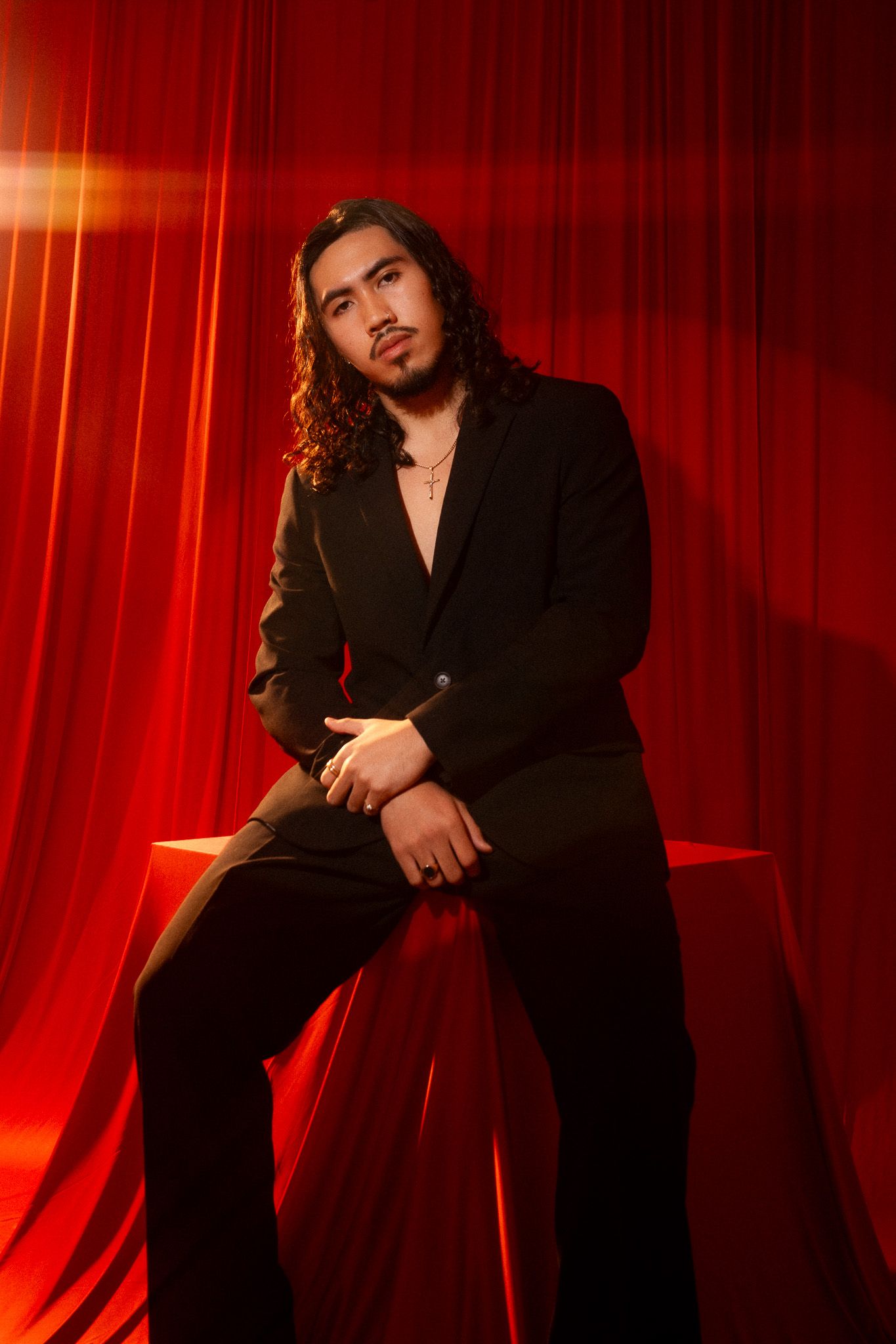 Stylish portrait against red backdrop shows figure in black suit with long wavy hair in dramatic lighting.