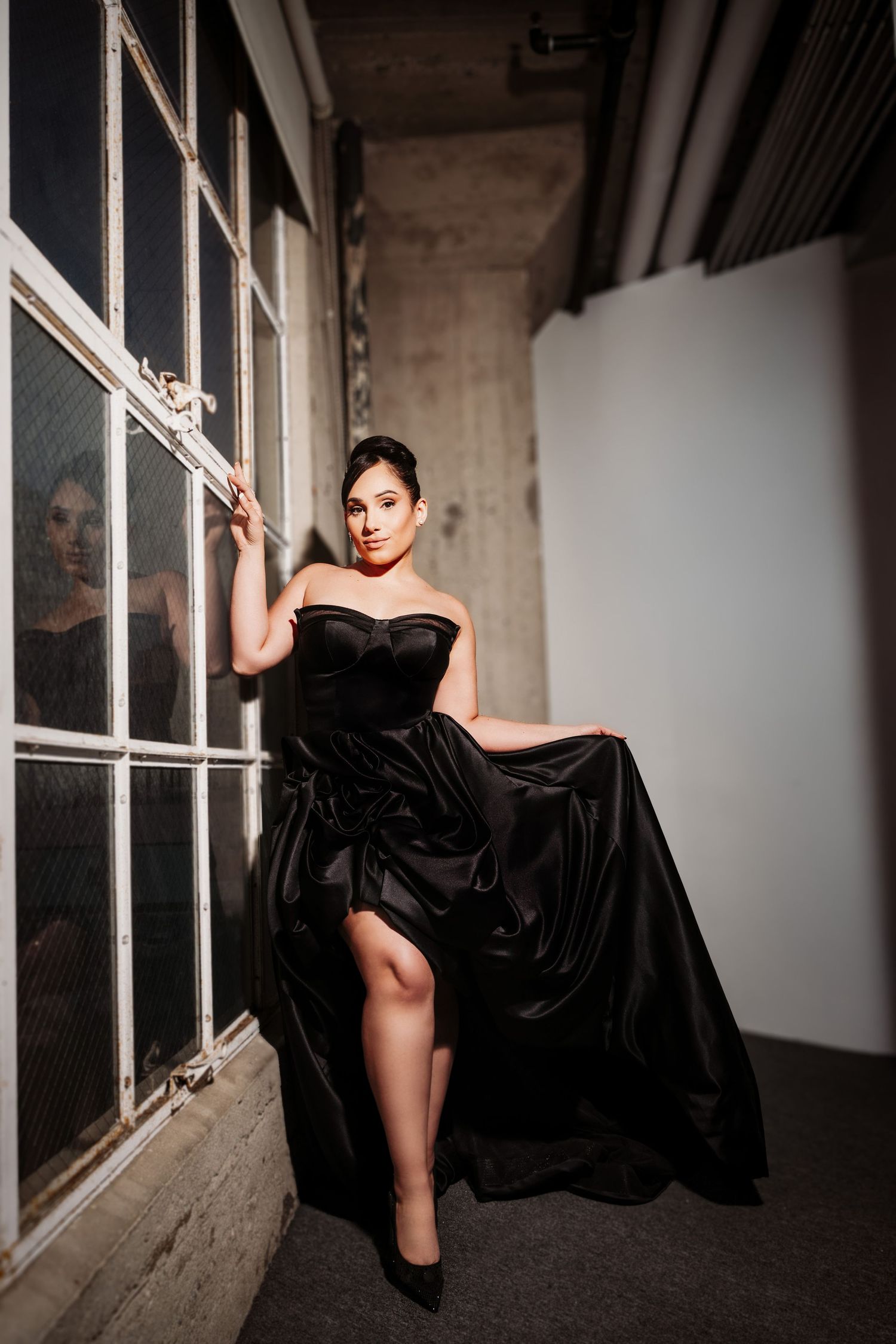 The person poses elegantly in a strapless black velvet gown next to tall industrial windows in a moody setting.