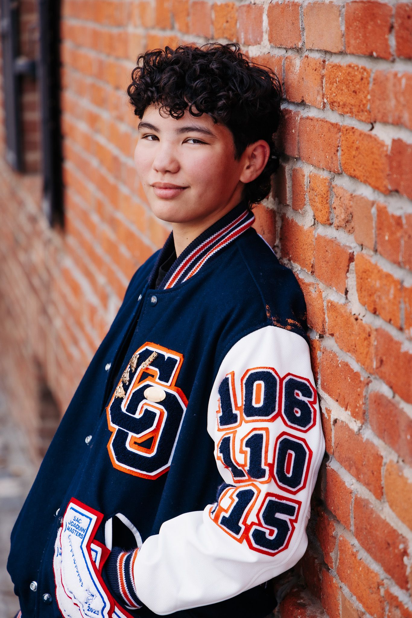 A person wearing a blue and white varsity letter jacket leans against a brick wall in a contemplative pose.